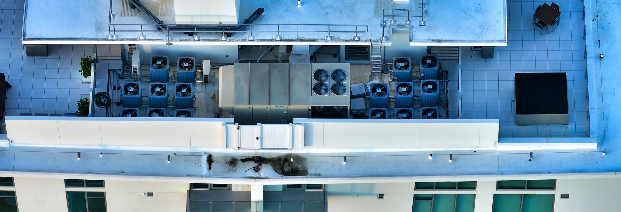 The image shows an aerial view of a rooftop space featuring multiple air conditioning units, a large mechanical unit, and a small sitting area with a table and chairs. The area is tiled with a light-colored surface and includes some greenery.