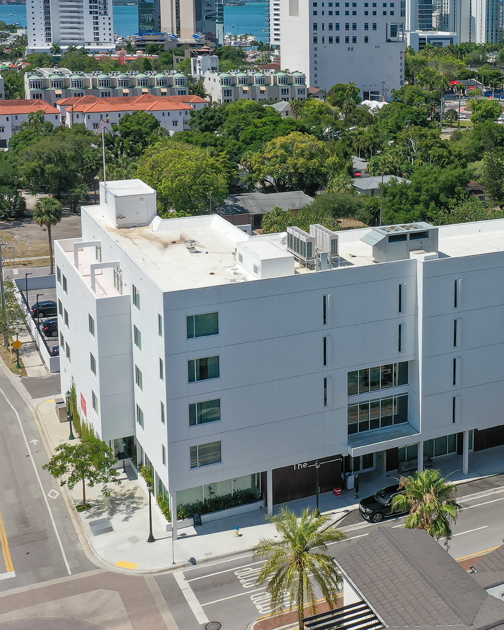 An aerial view of a modern white building located at a street corner in an urban area, featuring large windows and a contemporary design. Surrounding greenery and other buildings are visible, with a glimpse of the water in the background.