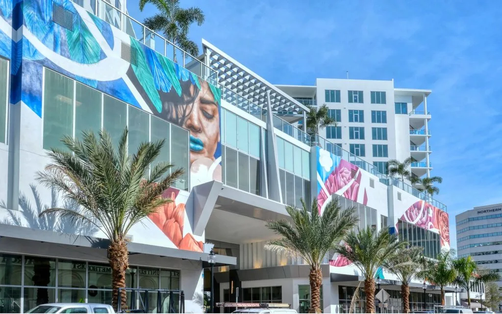 This image showcases a modern building adorned with colorful murals depicting various designs and faces. Palm trees are planted in front, enhancing the tropical aesthetic under a clear blue sky.