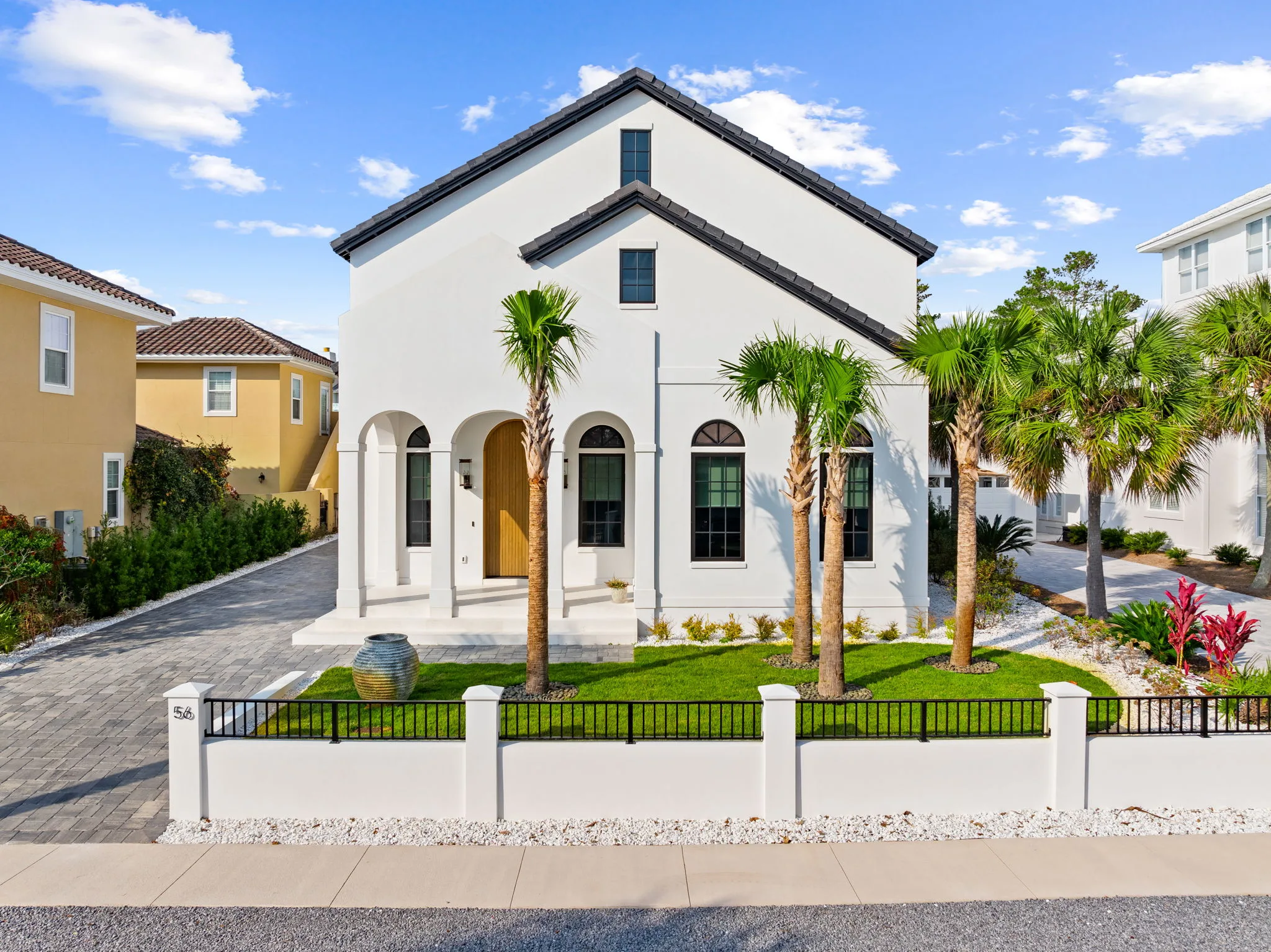 A white two-storey home with arched windows behind a low wall, palms and a green lawn.