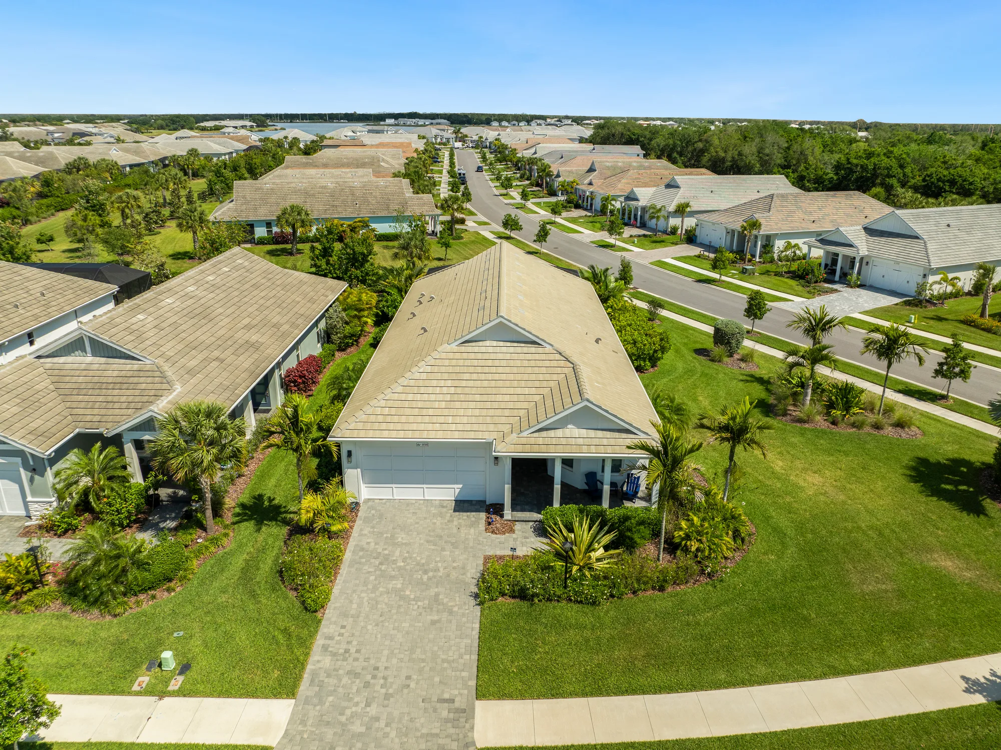 The image shows an aerial view of a suburban neighborhood featuring single-family homes with flat roofs and manicured lawns. Palm trees and landscaped gardens are visible, illustrating a tropical setting.