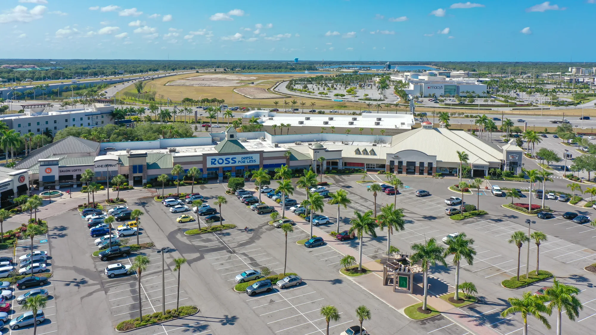 An aerial view of a shopping center featuring a Ross Dress for Less store and various other retail outlets. The surrounding parking lot is partially filled with cars, and palm trees line the area, set against a clear blue sky.