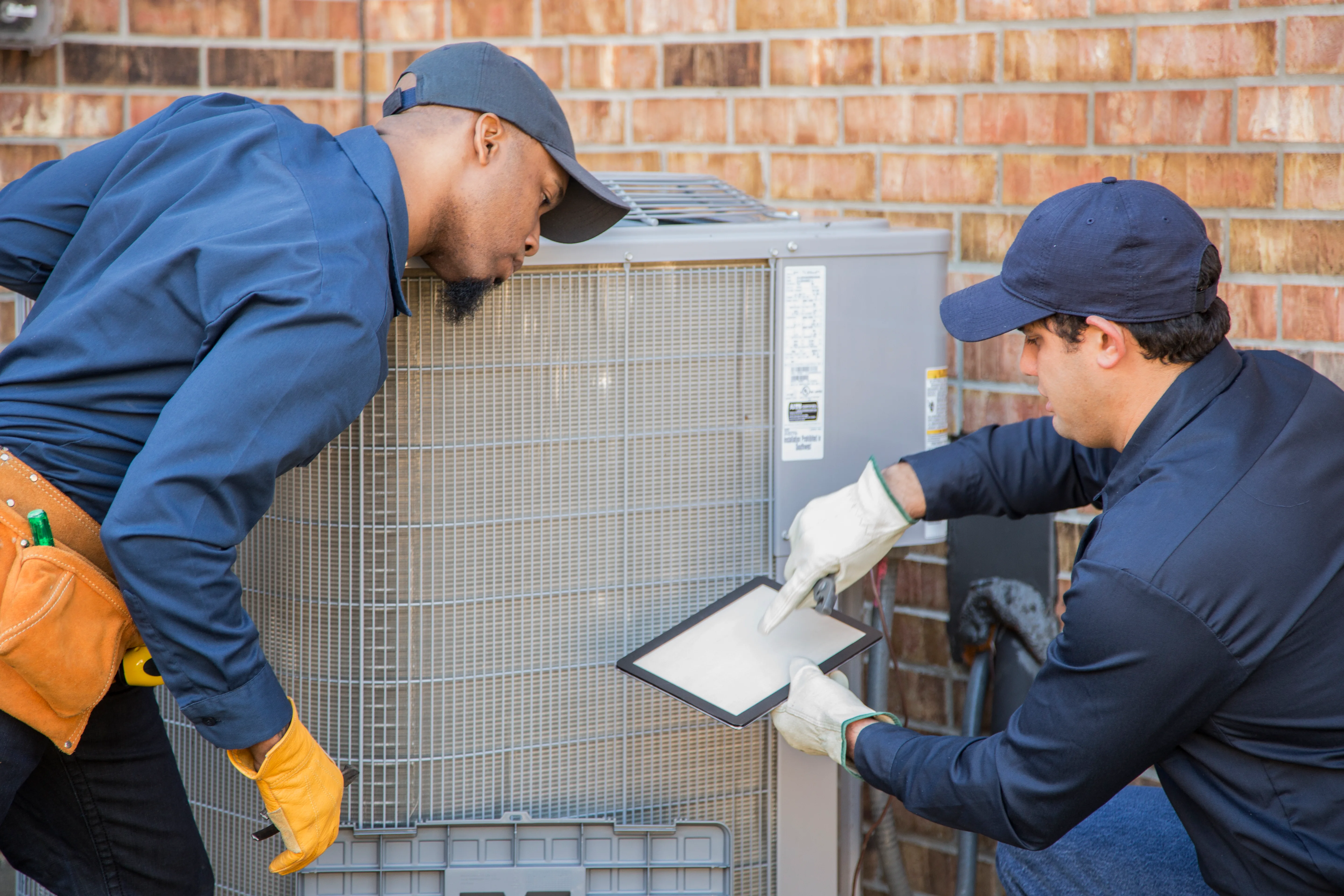 Two HVAC technicians are inspecting and servicing an outdoor air conditioning unit. One technician is looking closely at the unit while the other points to a device or document on a tablet.