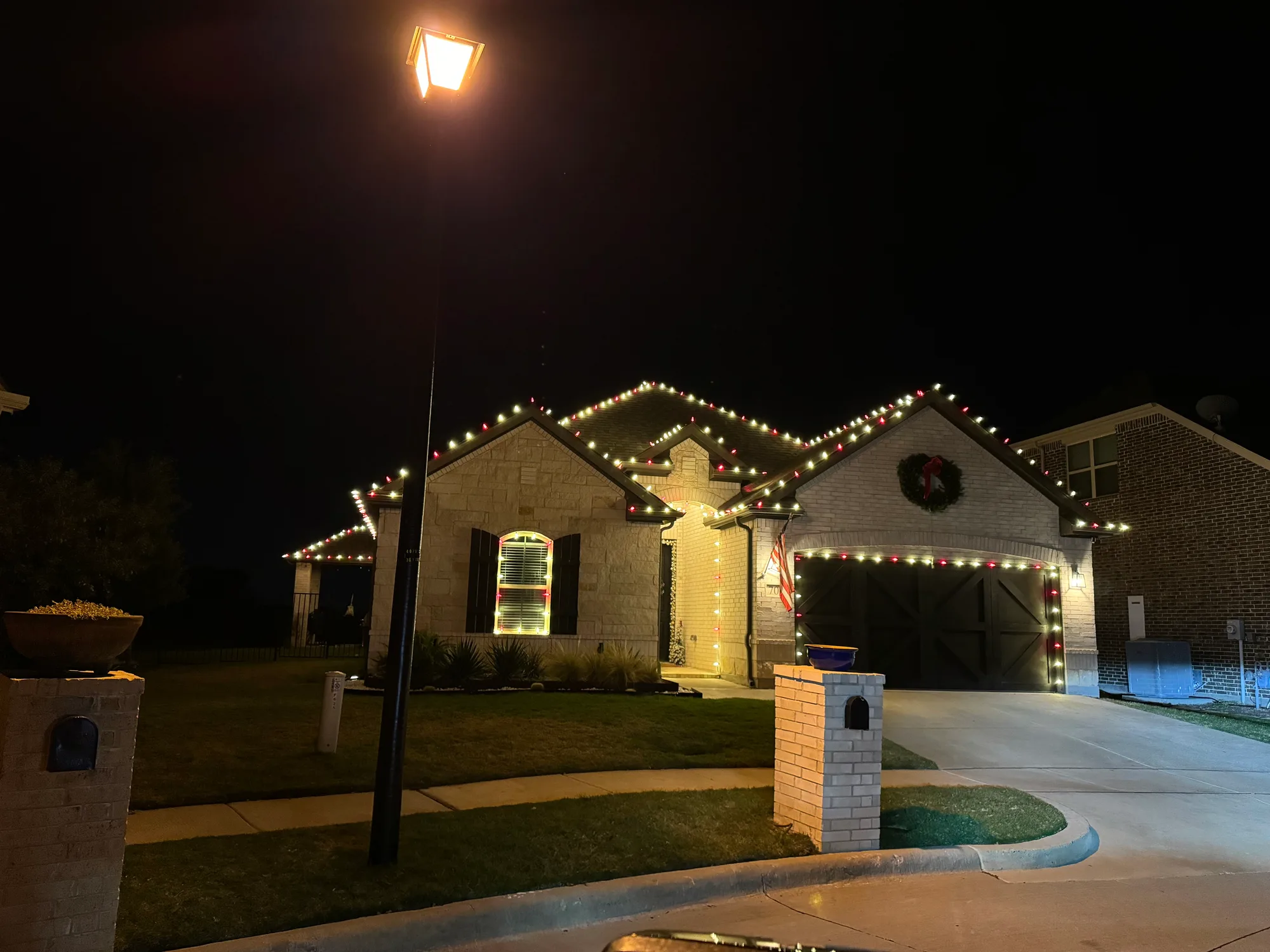 Stone-front home lit in alternating red and warm white, with the garage door outlined and a lit wreath mounted above it.