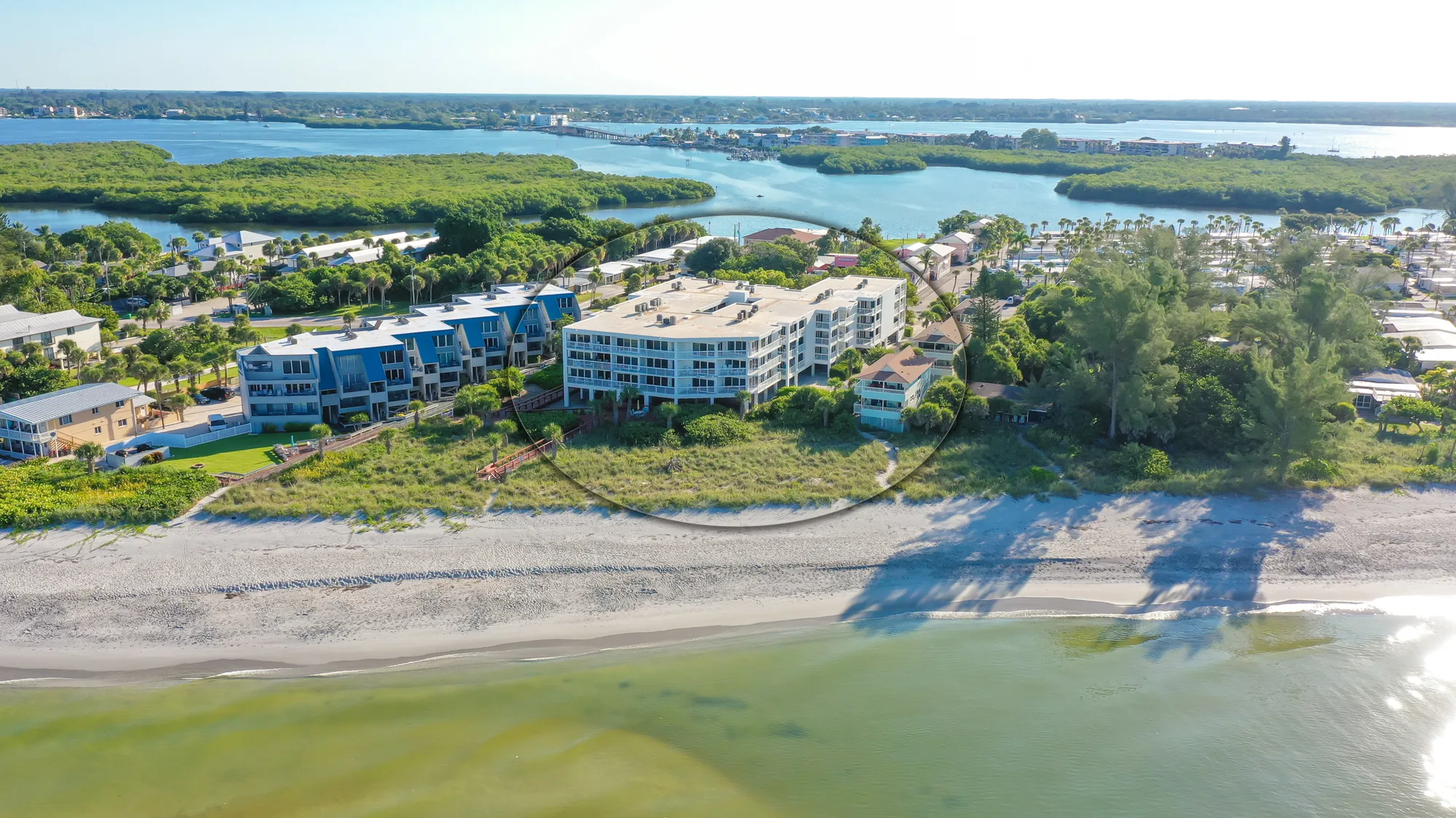An aerial view of a coastal resort situated along a sandy beach, featuring several buildings and lush greenery. The image captures the calm waters of a bay with mangroves in the background, highlighting the serene beach setting.