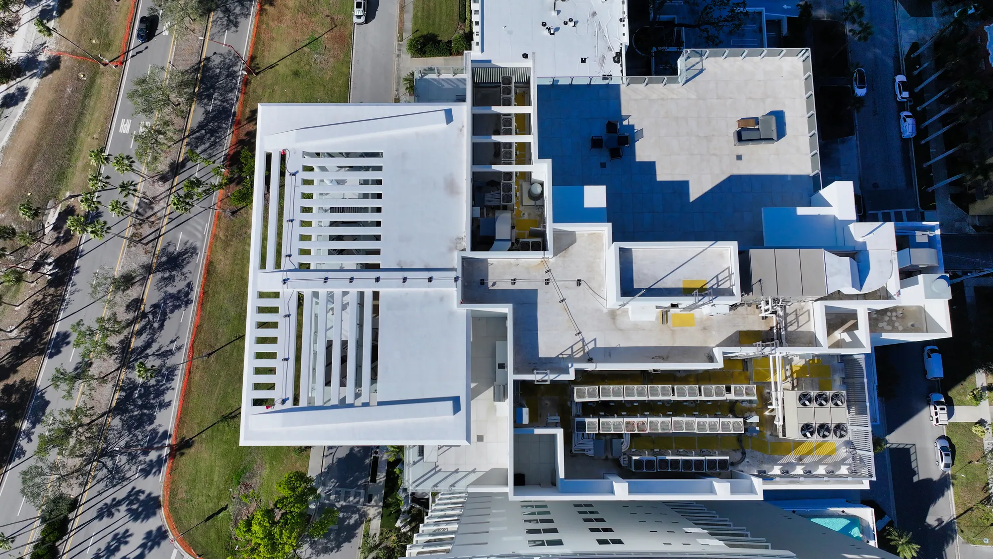 An aerial perspective of a modern building's rooftop, showcasing a layout with outdoor lounge areas, air conditioning units, and surrounding streets lined with palm trees. The image also reveals the geometric patterns and materials used in the building's design.