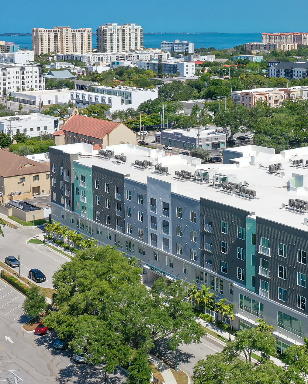 The image showcases a modern multi-story apartment building with a sleek exterior featuring a mix of dark and light colors. In the background, a city skyline with several high-rise buildings is visible, along with a hint of blue water, indicating proximity to the coast.