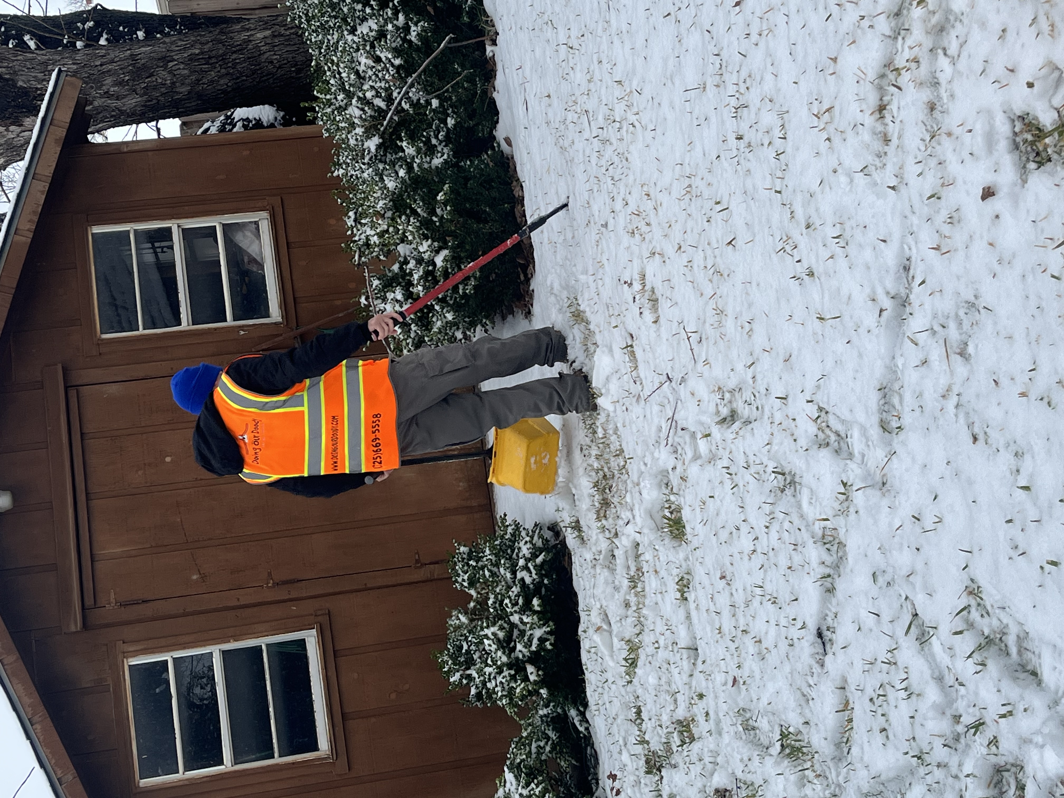 Worker in reflective safety vest shoveling snow in front of a wooden shed