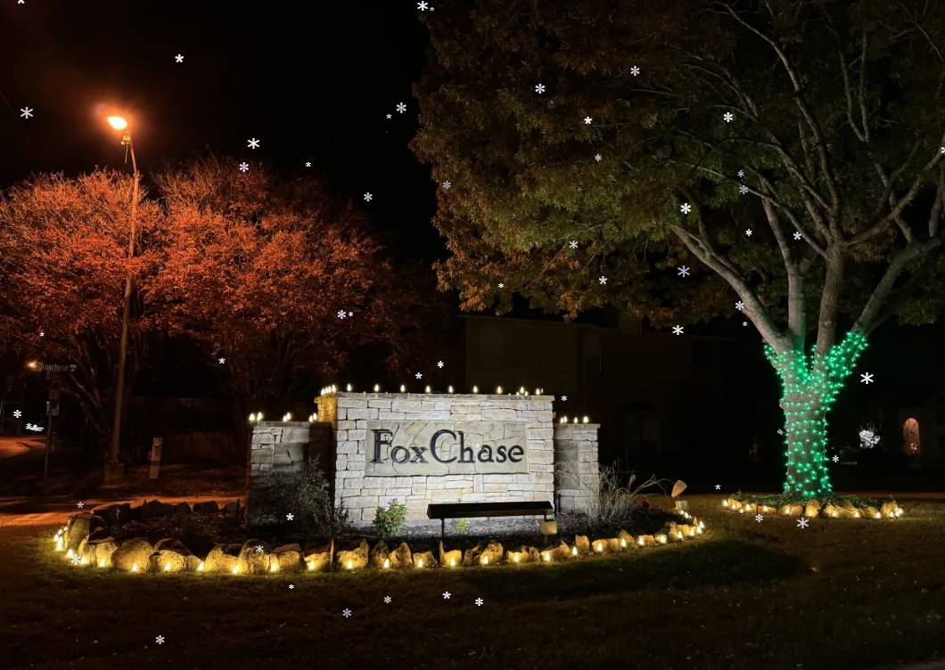 Fox Chase neighbourhood entrance sign lit at night, with warm white bulbs along the top of the stone wall, ground lights around the planting bed and a tree trunk wrapped in green lights.