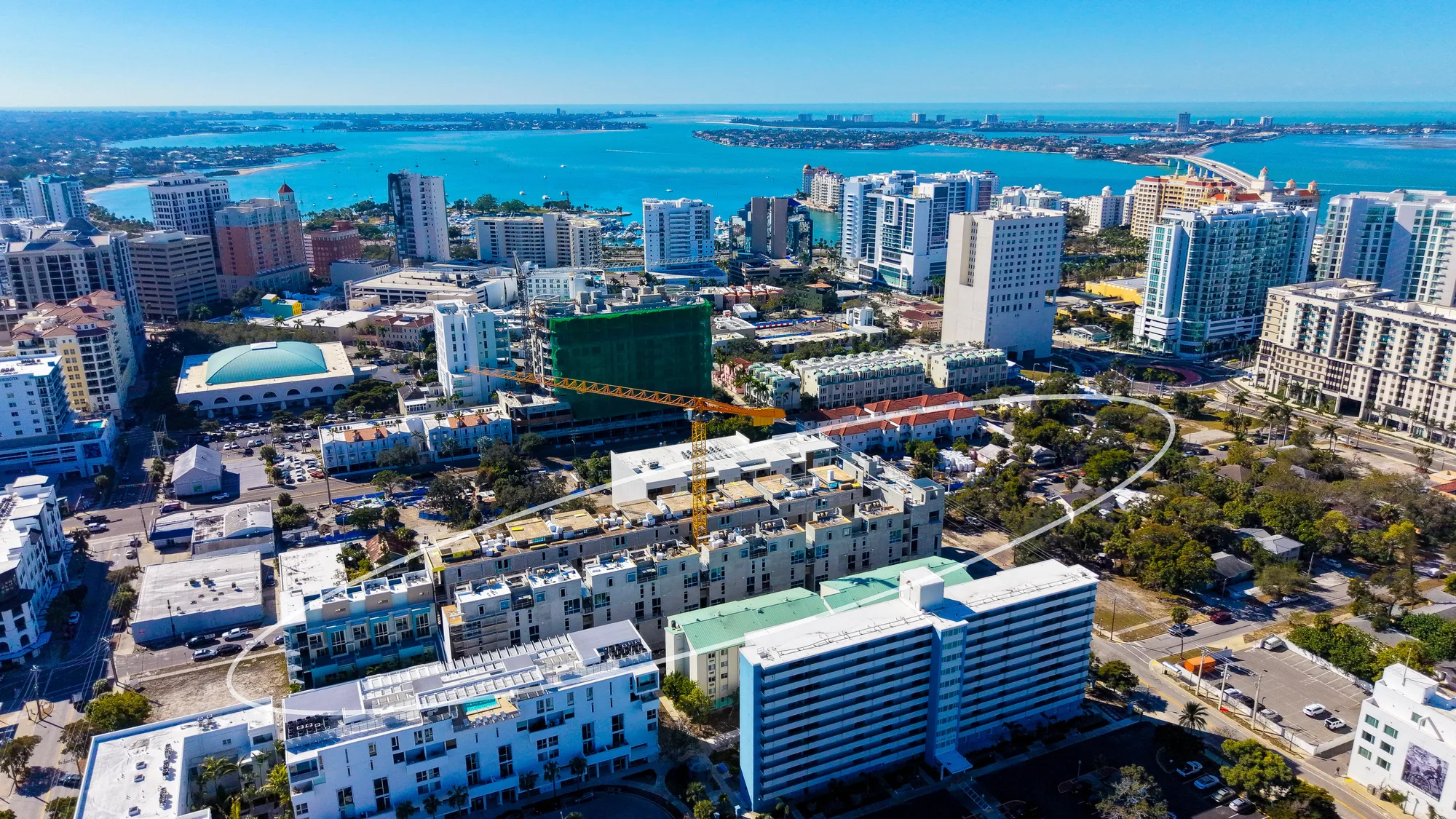 This aerial image showcases a bustling coastal city featuring high-rise buildings, ongoing construction with a crane, and a panoramic view of the waterfront. The vibrant skyline is complemented by clear blue skies and the sparkling waters of the bay.