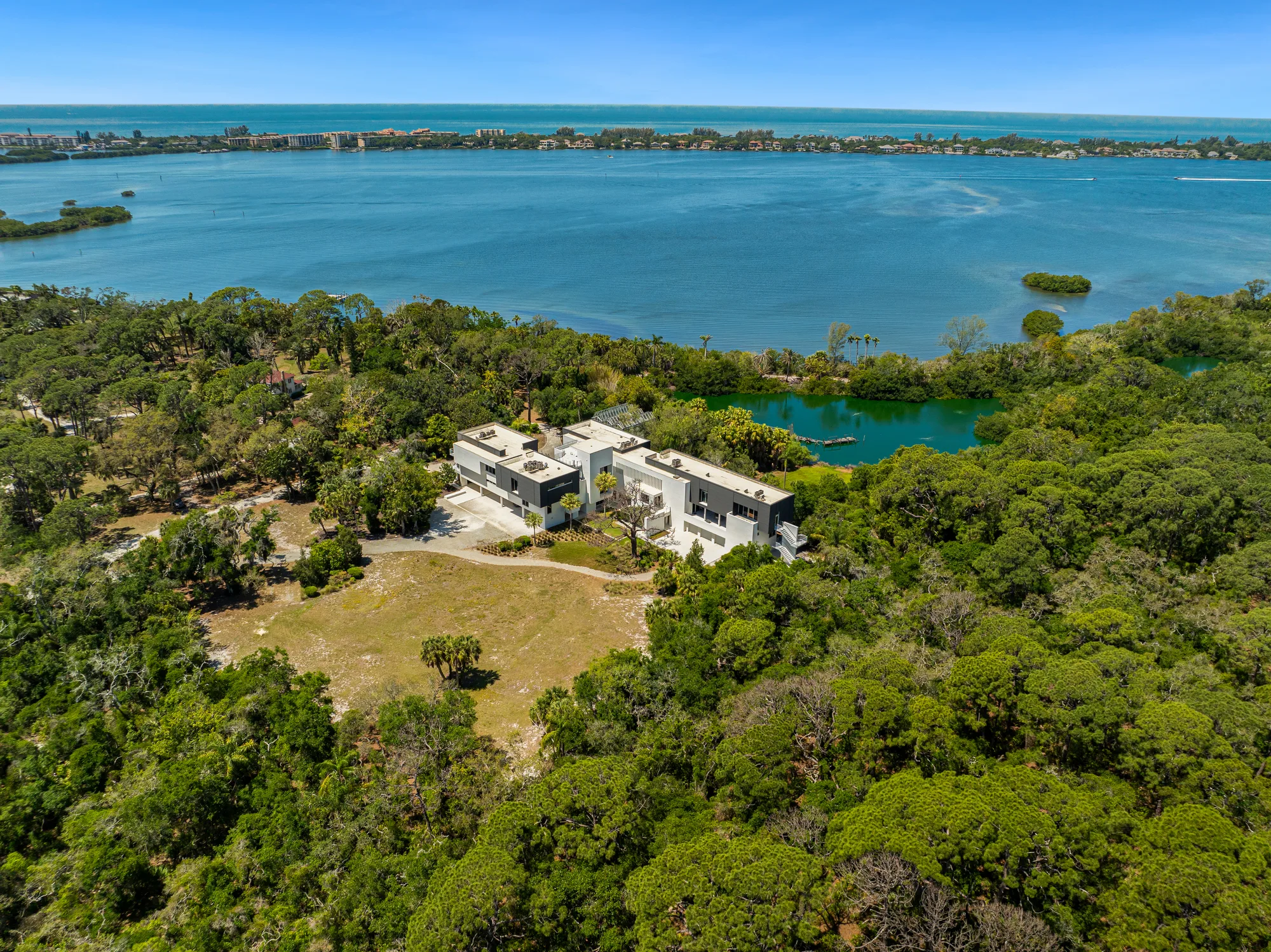 An aerial view of a modern two-story house nestled among lush greenery, with a scenic waterfront and distant coastal views in the background. The property features landscaped areas and is situated near a lagoon, showcasing a blend of natural beauty and contemporary architecture.