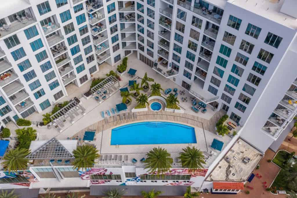 An aerial view of a modern apartment complex featuring a central swimming pool surrounded by lounge chairs and tropical landscaping. The building showcases multiple balconies with seating areas, emphasizing a contemporary architectural design.