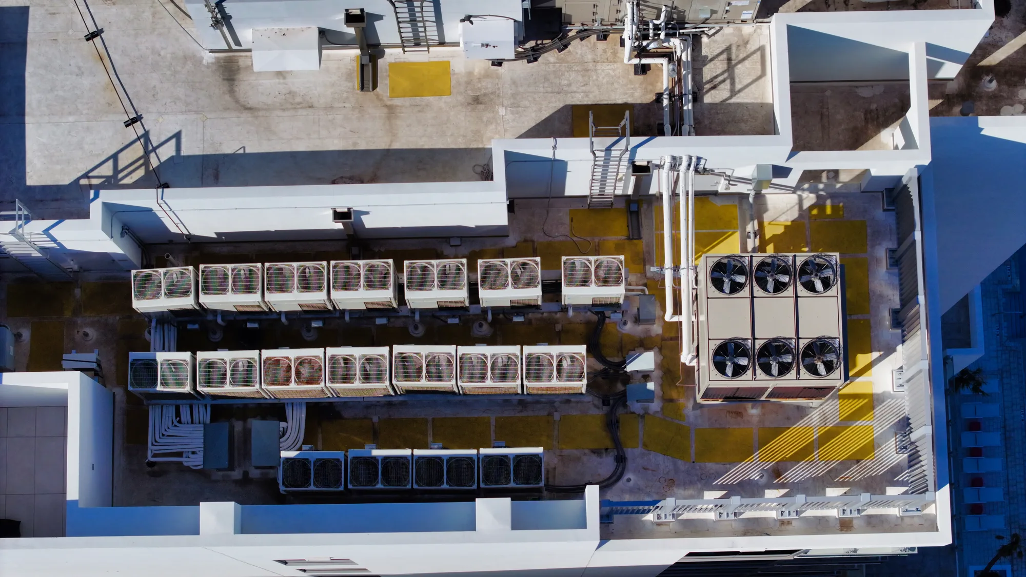 An aerial view of a rooftop featuring multiple HVAC units arranged in lines. The image showcases a combination of air conditioning units and large cooling fans, along with visible piping and a concrete surface.