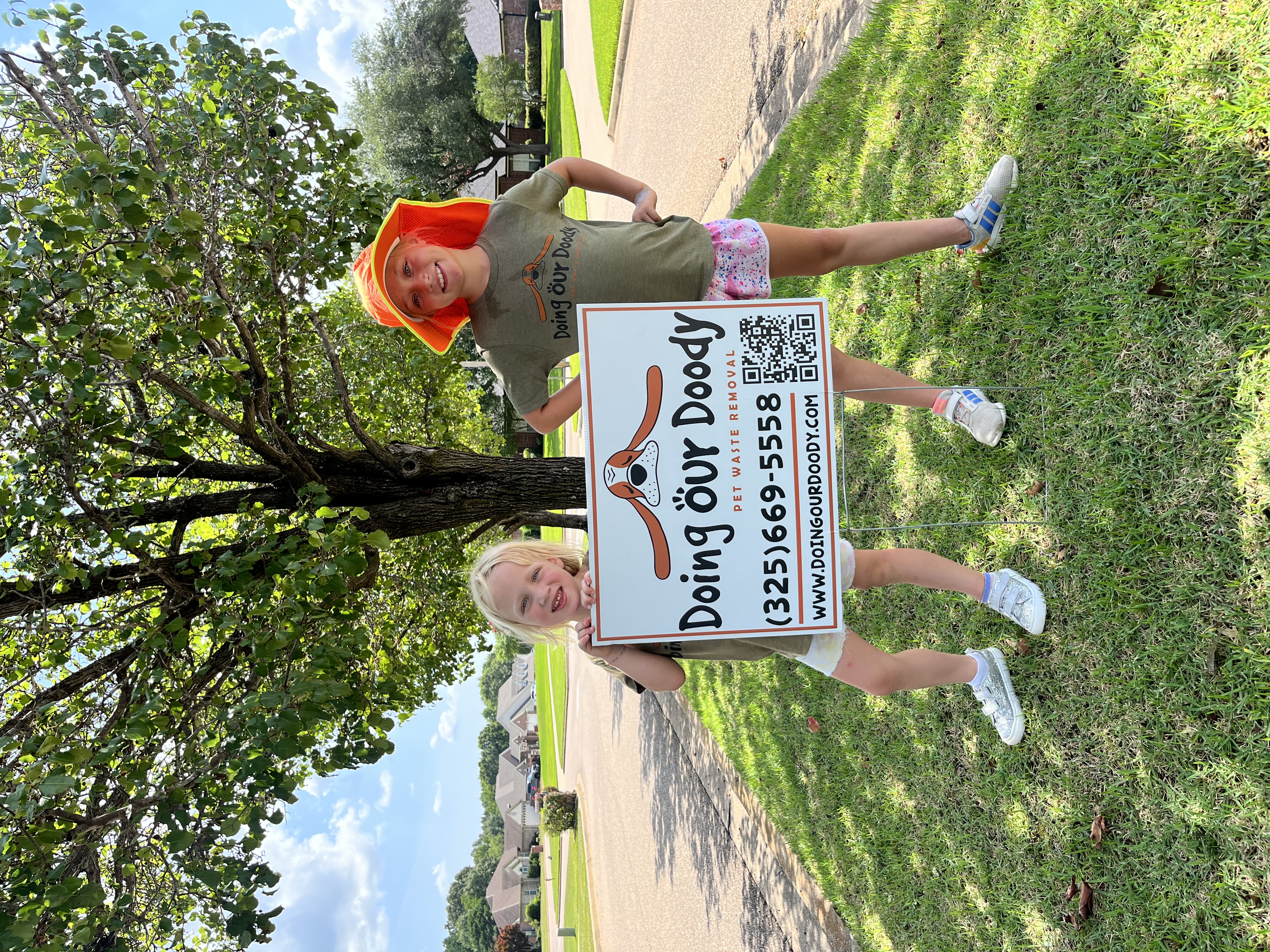 Two young girls smiling in front of a Doing Our Doody pet waste removal sign