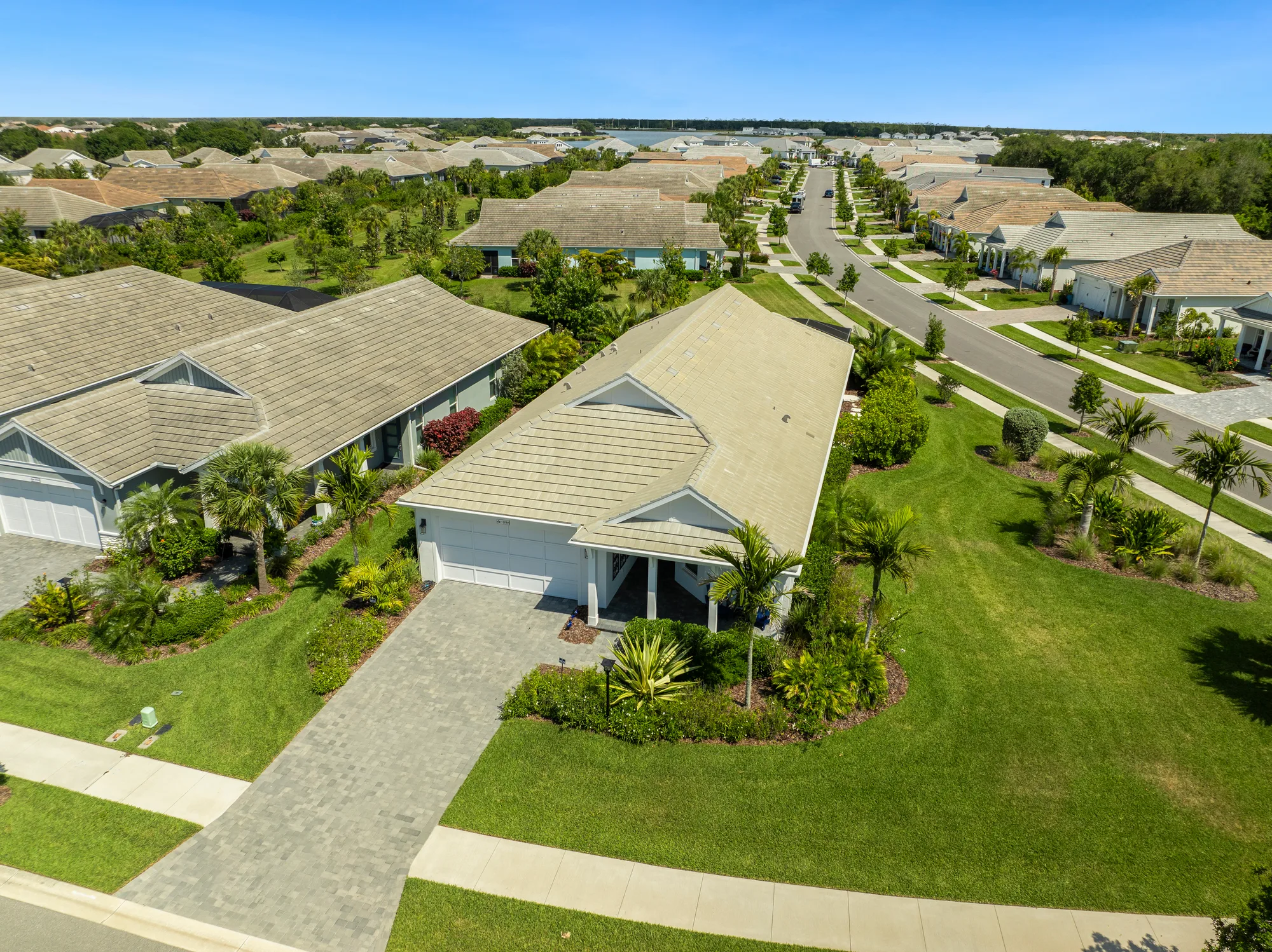 An aerial view of a suburban neighborhood featuring well-maintained houses, neatly landscaped yards, and paved driveways. The scene includes a wide street lined with homes, surrounded by lush greenery and palm trees.