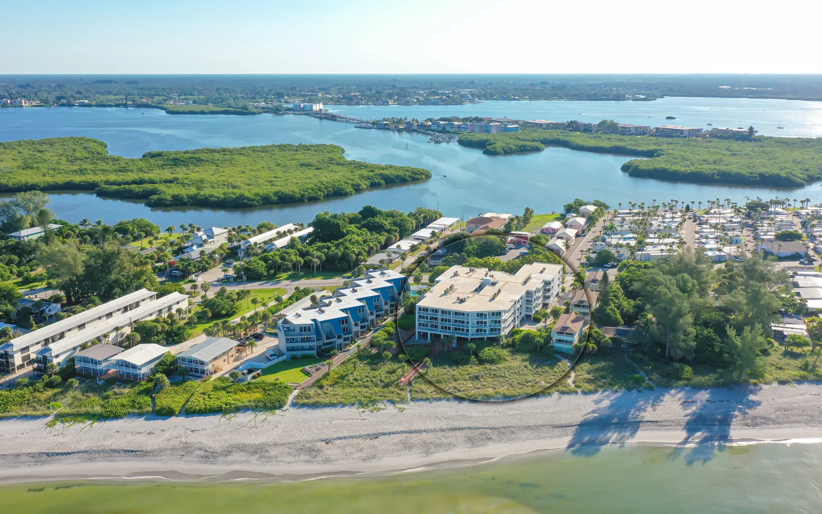 An aerial view of a coastal area featuring residential buildings along the beach, lush greenery, and waterways. The scene captures a blend of waterfront properties and serene natural landscapes.