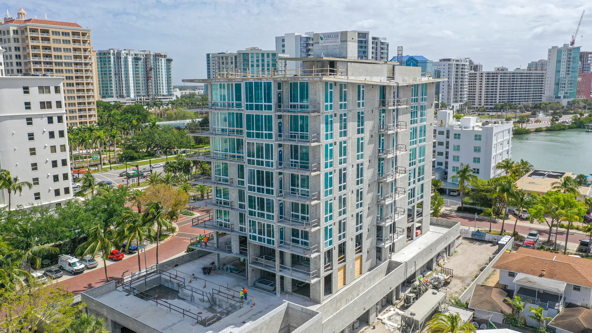An aerial view of a modern high-rise building under construction, featuring multiple glass windows and a concrete structure. Surrounding the building are palm trees, roads, and other urban developments, showcasing a vibrant city environment.