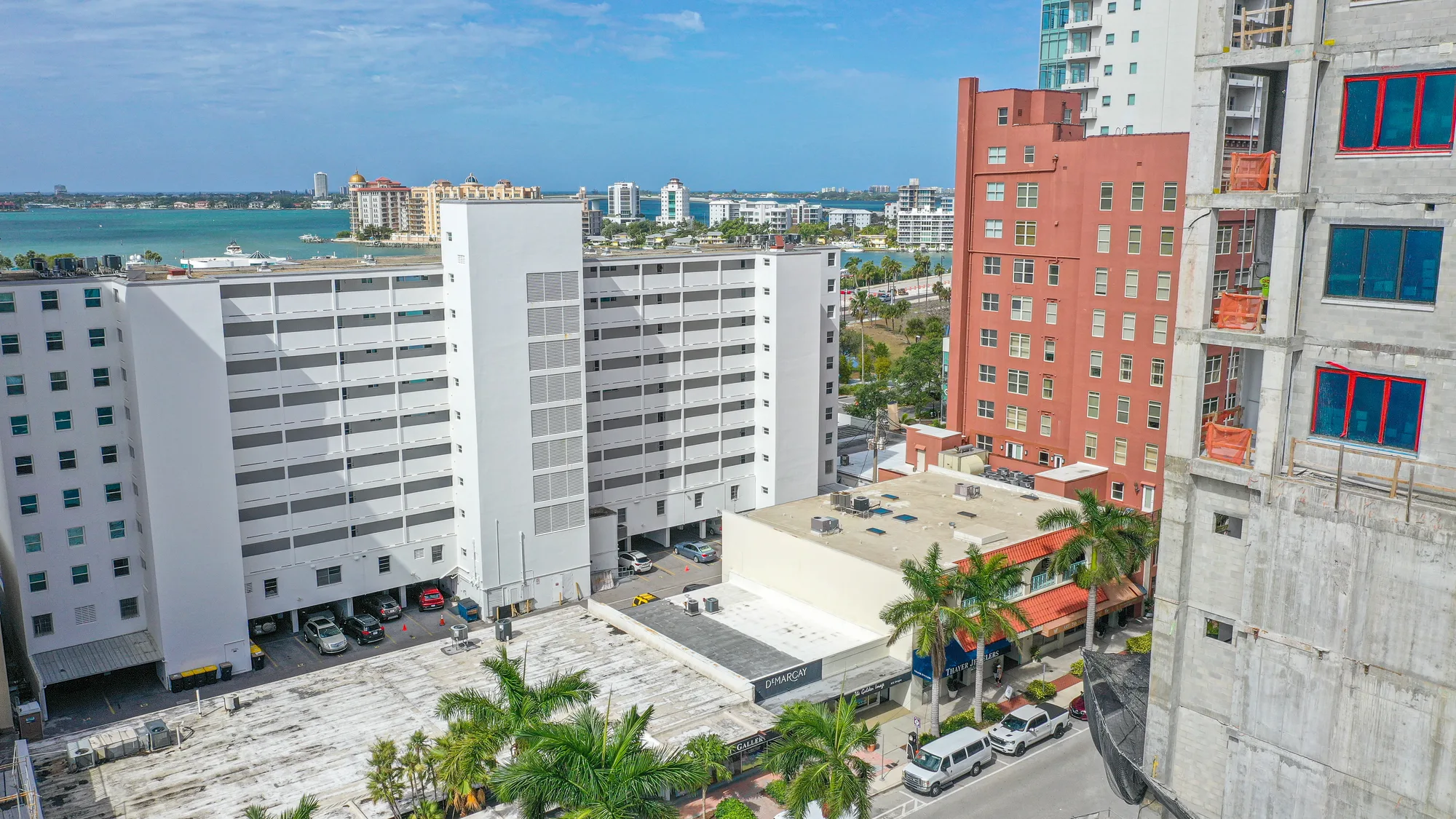 The image showcases a modern urban setting featuring a parking structure and multiple residential buildings in the background, alongside a waterfront view. Palm trees populate the foreground, contributing to the tropical atmosphere.