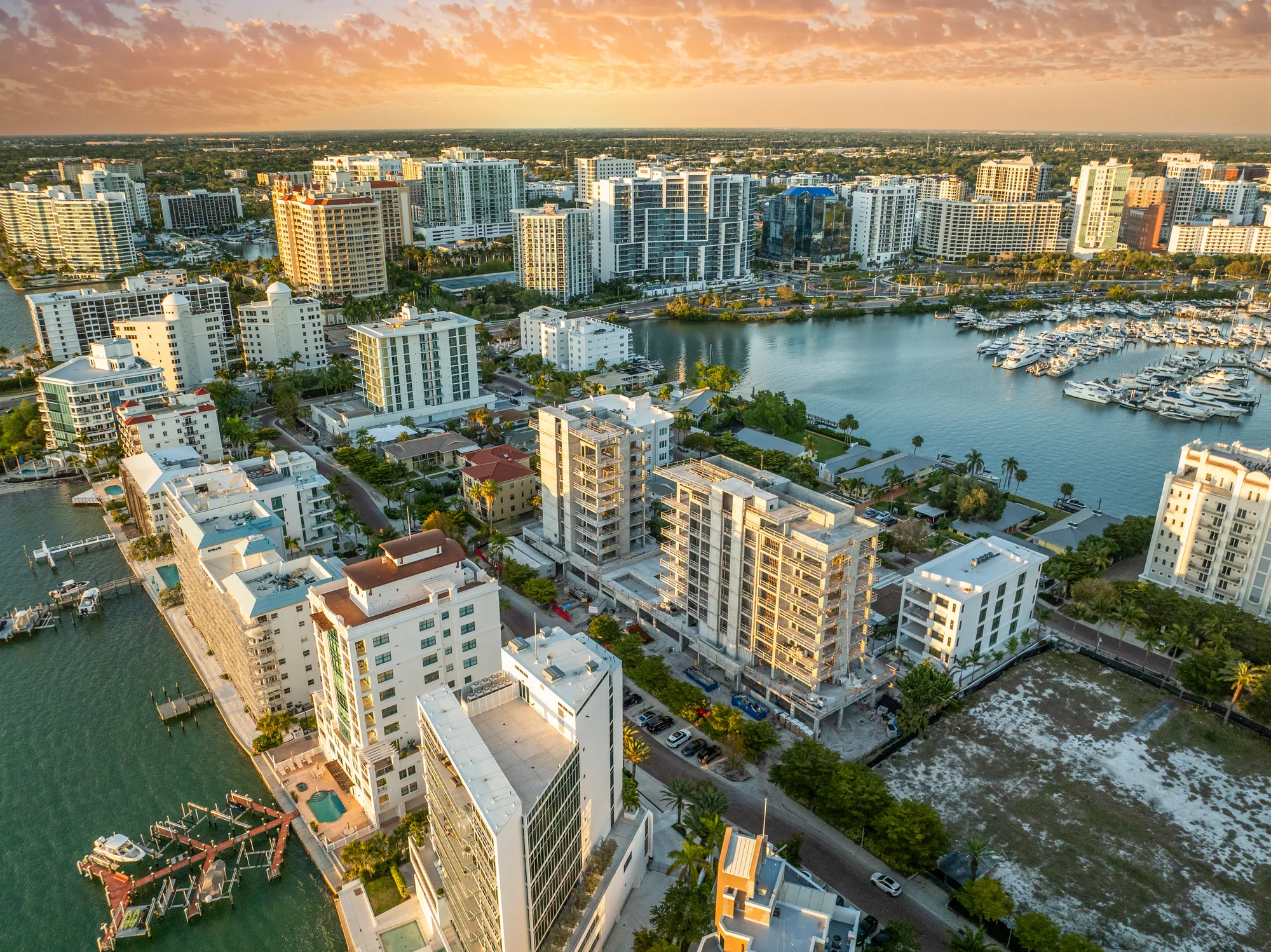 This aerial image showcases a modern urban waterfront area with high-rise buildings, marinas, and lush greenery. The scene is illuminated by a warm sunset, creating a picturesque view of the city alongside a calm body of water.