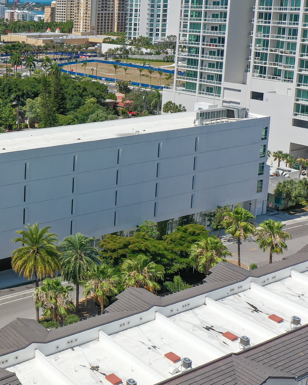 The image showcases a contemporary building with a minimalist white facade surrounded by lush greenery and palm trees. In the background, a skyline with various high-rise buildings can be seen, highlighting an urban setting.