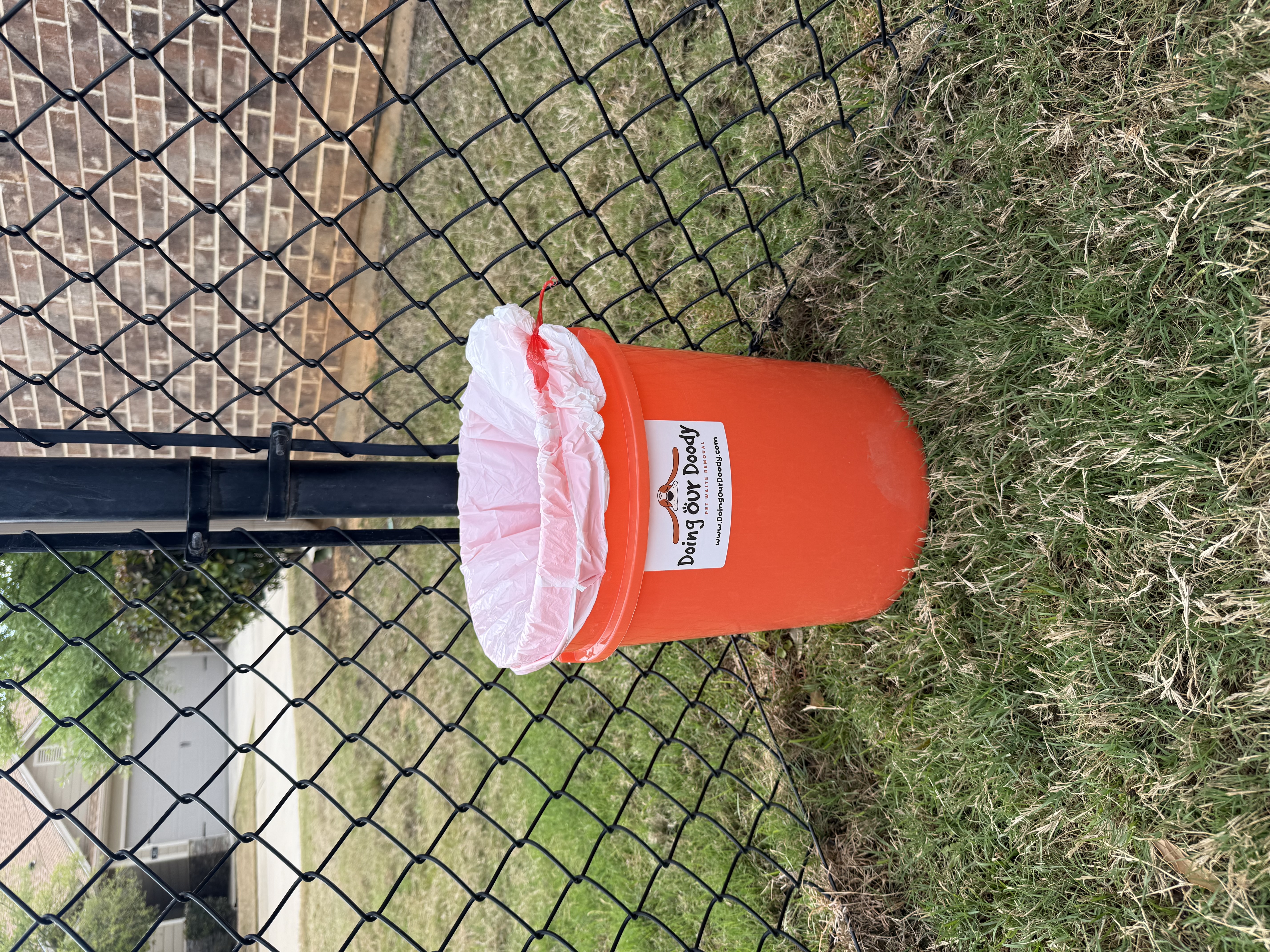 Orange Doing Our Doody pet waste disposal bin next to a black chain-link fence