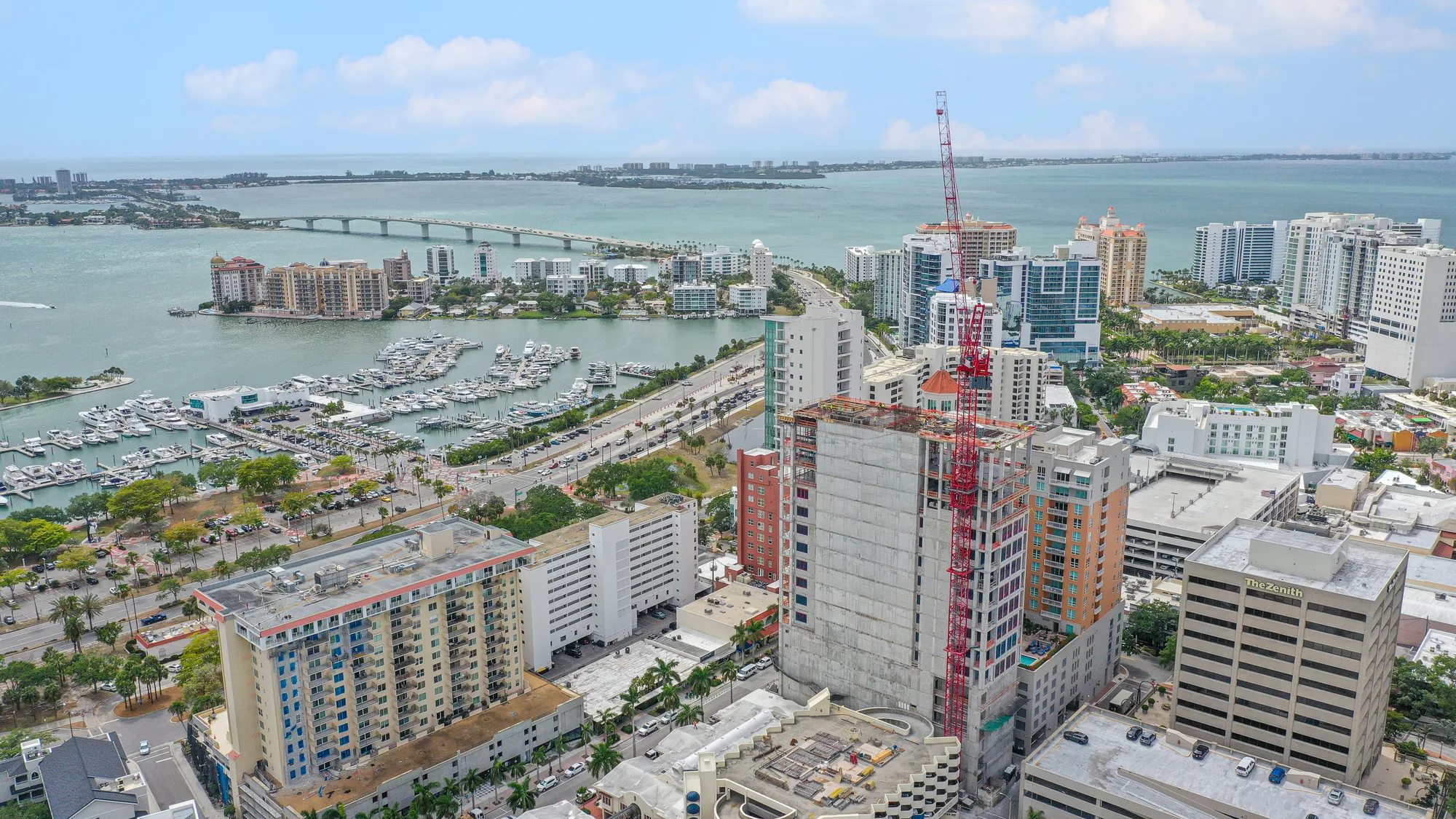 This aerial image captures the Miami skyline featuring a mix of residential and commercial buildings, with a marina and bridge visible in the background. Construction activity is evident with a crane on a building site, showcasing the city's ongoing development.