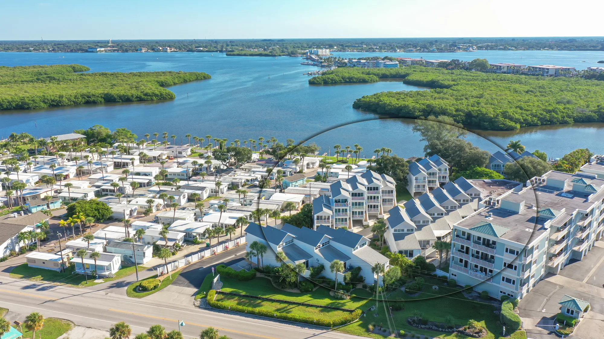 This aerial image captures a coastal community with a mix of residential buildings and mobile homes adjacent to a serene waterway. The surrounding lush greenery and blue water highlight the area's natural beauty.