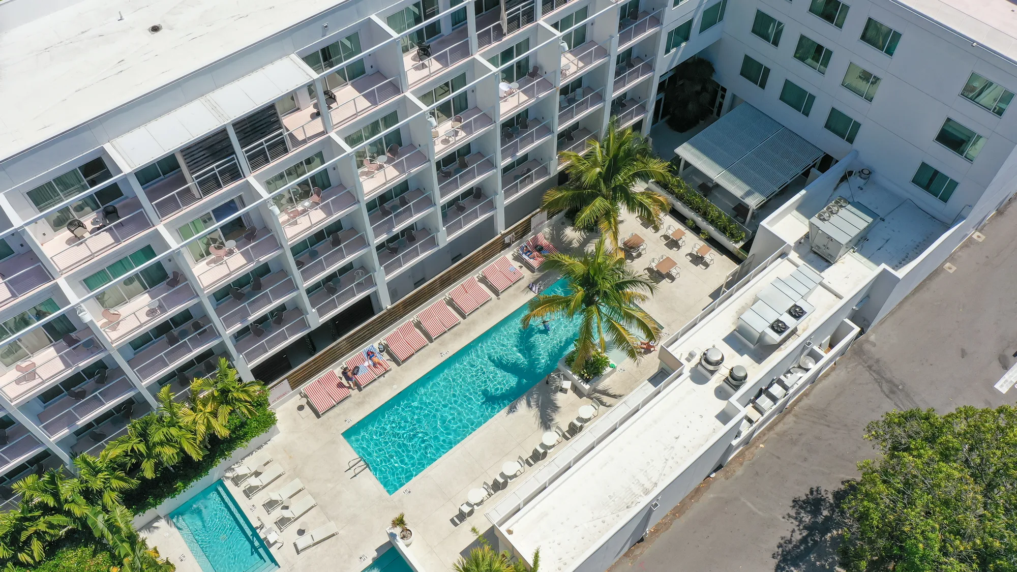 The image showcases an aerial view of a hotel complex featuring a swimming pool surrounded by lounge chairs, palm trees, and the building's balconies. The pool area is well-maintained, with guests relaxing by the water under the bright sun.
