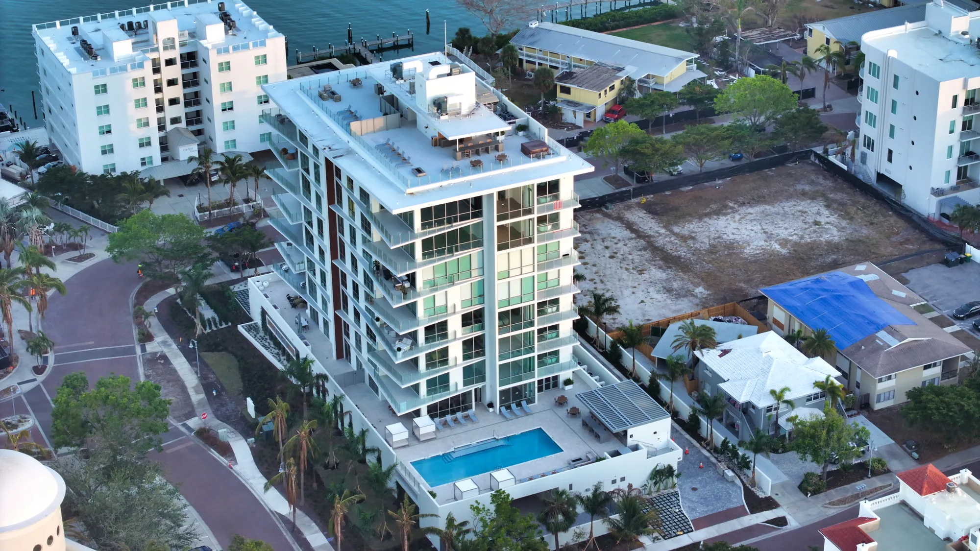 An aerial view of a contemporary apartment building featuring multiple floors of glass balconies and a rooftop terrace, alongside a swimming pool at ground level. Surrounding the building are palm trees and a mix of residential properties, with some undeveloped land nearby.
