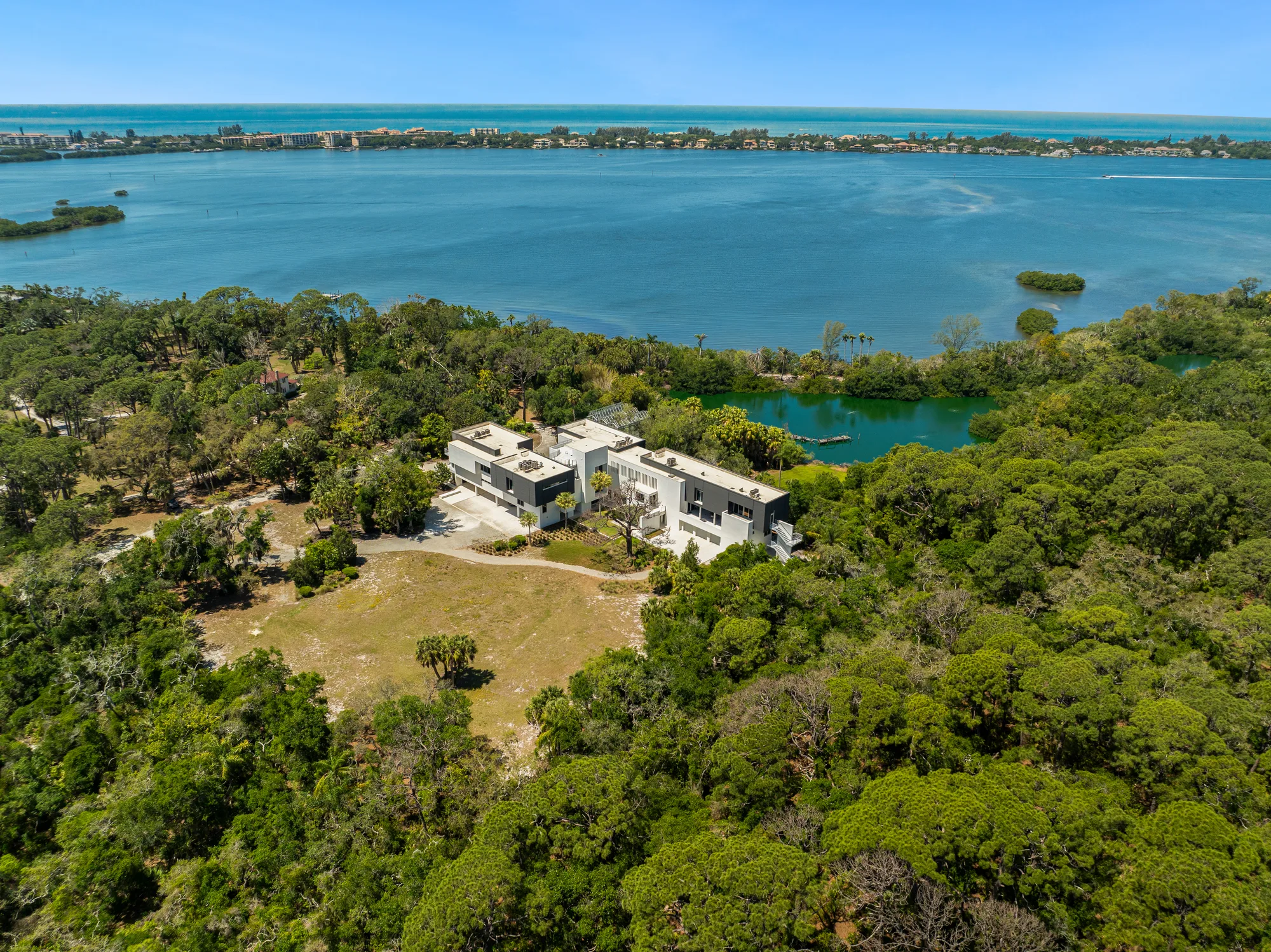 Aerial view of a contemporary two-story house surrounded by lush greenery, adjacent to a serene body of water. The landscape includes nearby trees and a glimpse of the coastline in the background.