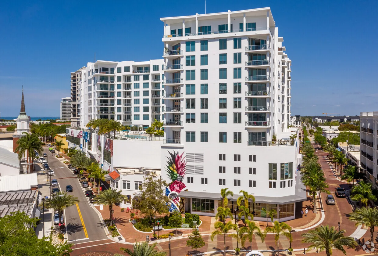Street-level view of The Mark, a white curved residential tower in downtown Sarasota, framed by palms.