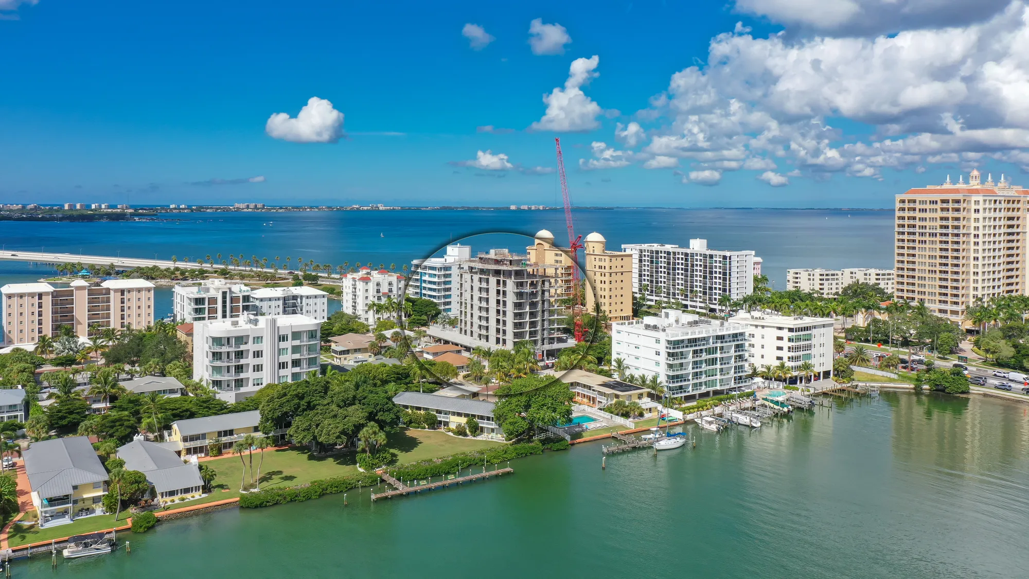 An aerial view of a coastal urban area featuring modern buildings alongside a serene blue harbor. The scene includes lush greenery, a marina with boats, and a clear sky with scattered clouds.