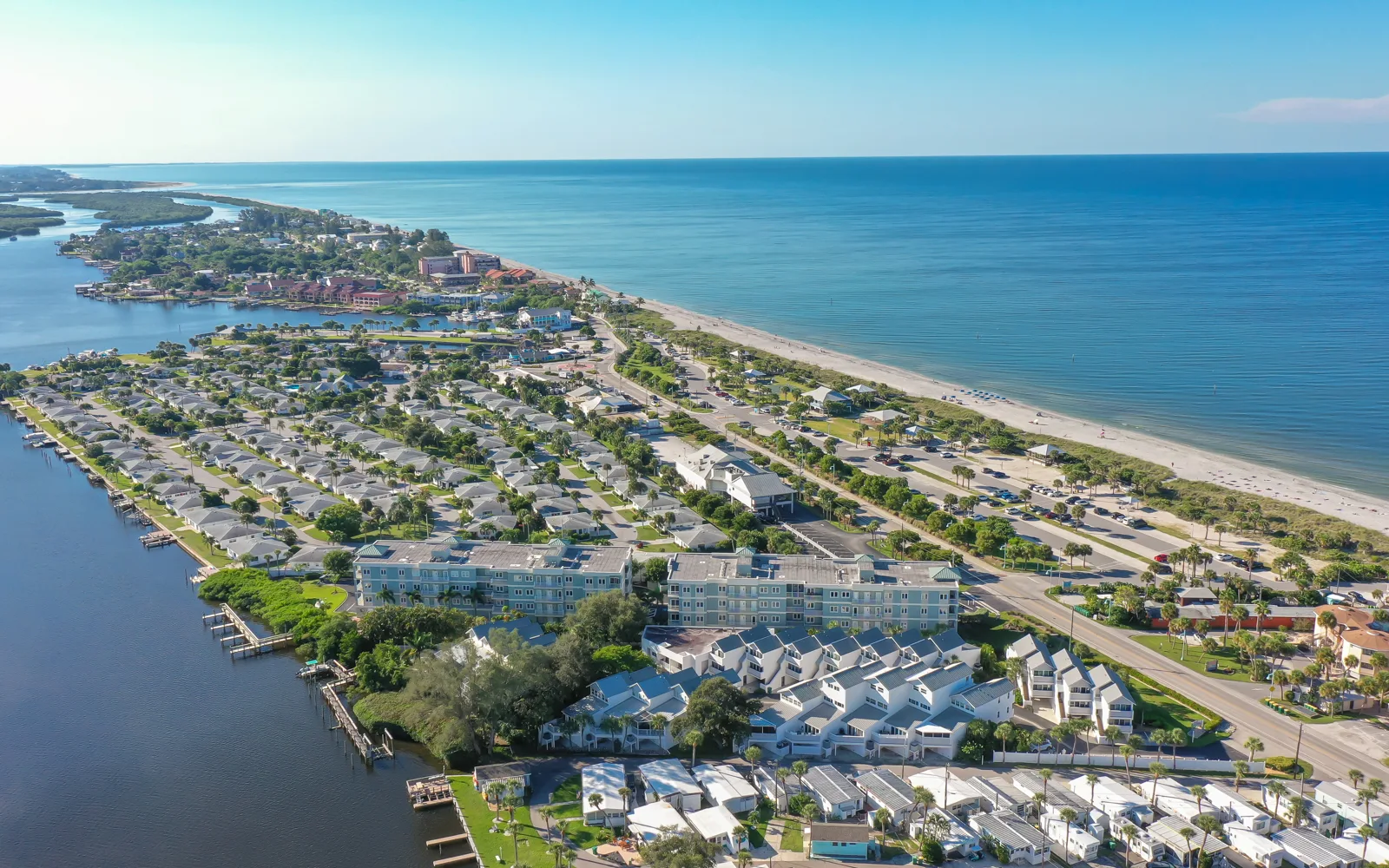 This aerial image captures a coastal community featuring residential buildings along a waterfront, with a sandy beach and the ocean visible in the distance. Lush greenery and roads are interspersed among the homes, creating a picturesque seaside landscape.