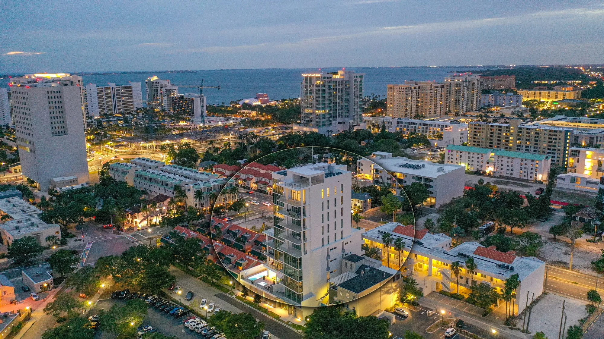 The image captures a vibrant aerial view of a coastal city at dusk, showcasing a modern high-rise building surrounded by a mix of residential and commercial structures. The scene is illuminated with city lights, highlighting the urban layout and proximity to the waterfront.