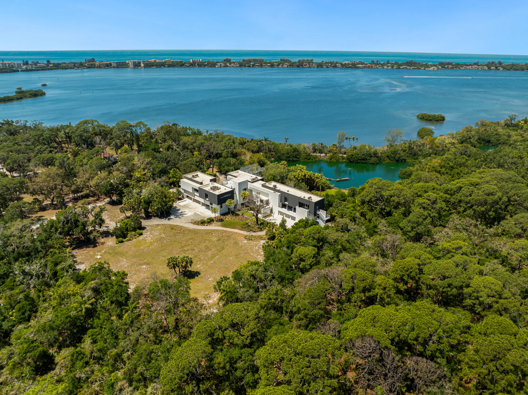 An aerial view of a contemporary waterfront home nestled among lush greenery, with a serene lake and coastal waters in the background. The property features landscaped grounds and a blend of natural surroundings.