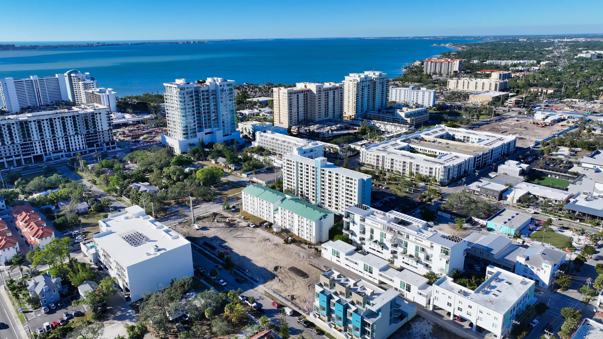 This aerial image captures a vibrant coastal cityscape featuring a mix of high-rise buildings, residential complexes, and green spaces. The view also includes a serene blue body of water in the background, showcasing the urban development against the coastal landscape.