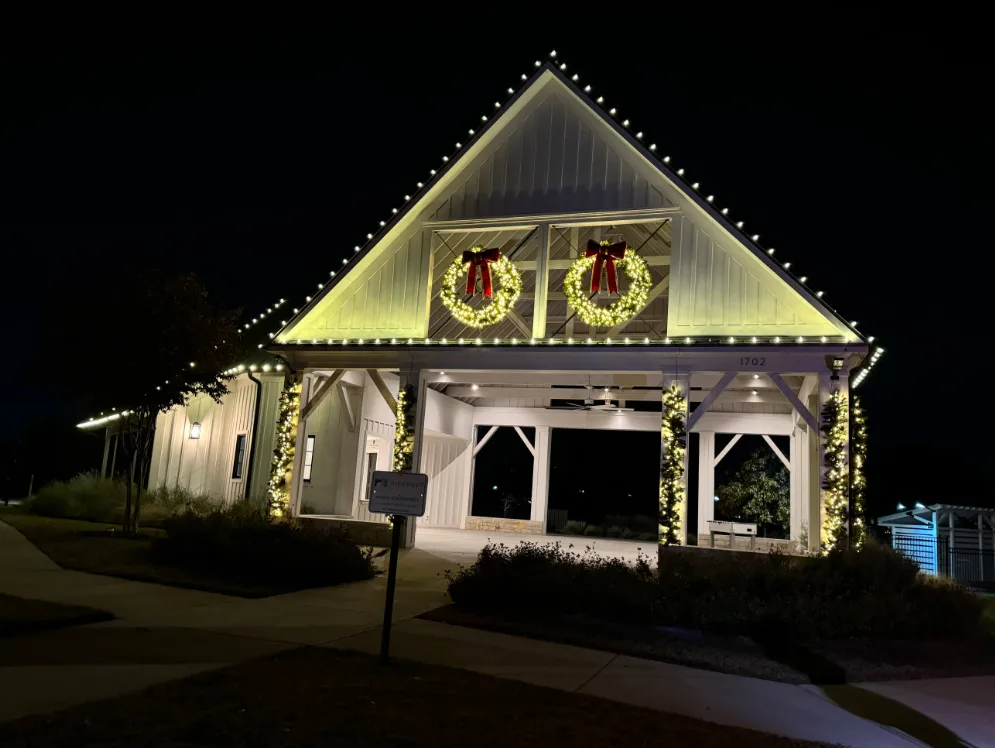 White community pavilion at night with warm white bulbs following the full gable roofline, two large lit wreaths with red bows on the gable, and garland wrapped up every column.