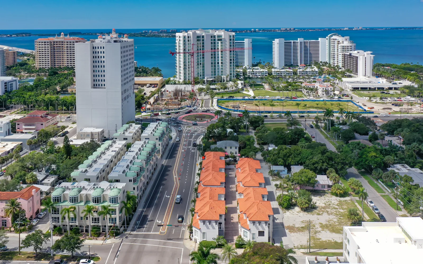 This aerial image showcases a bustling coastal area featuring a mix of high-rise buildings and low-rise residential structures, with a roundabout and palm trees visible. In the background, a calm body of water reflects the clear sky, enhancing the vibrant urban landscape.