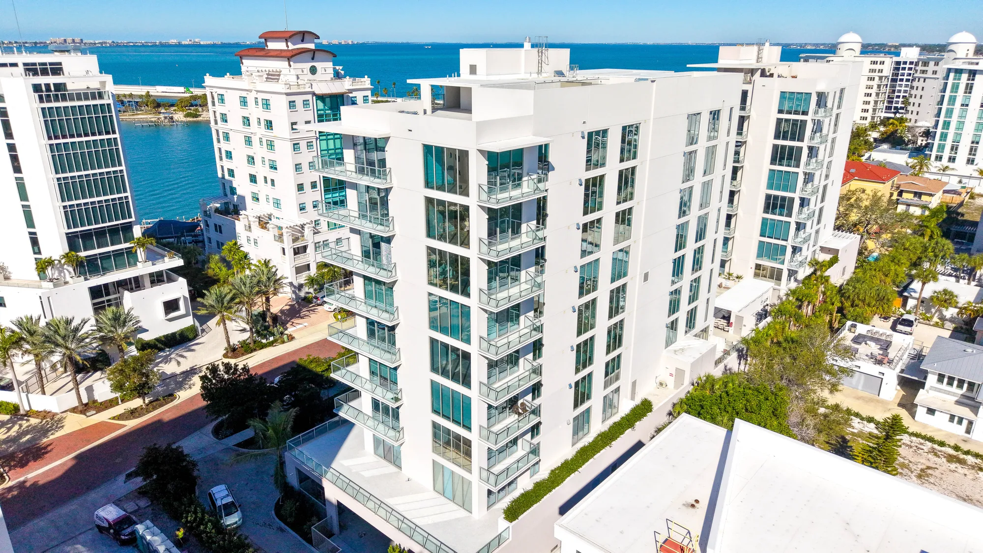 Aerial of The Collection Residences in Sarasota, a white multi-storey residential building among the downtown low-rise blocks, with the Gulf beyond.