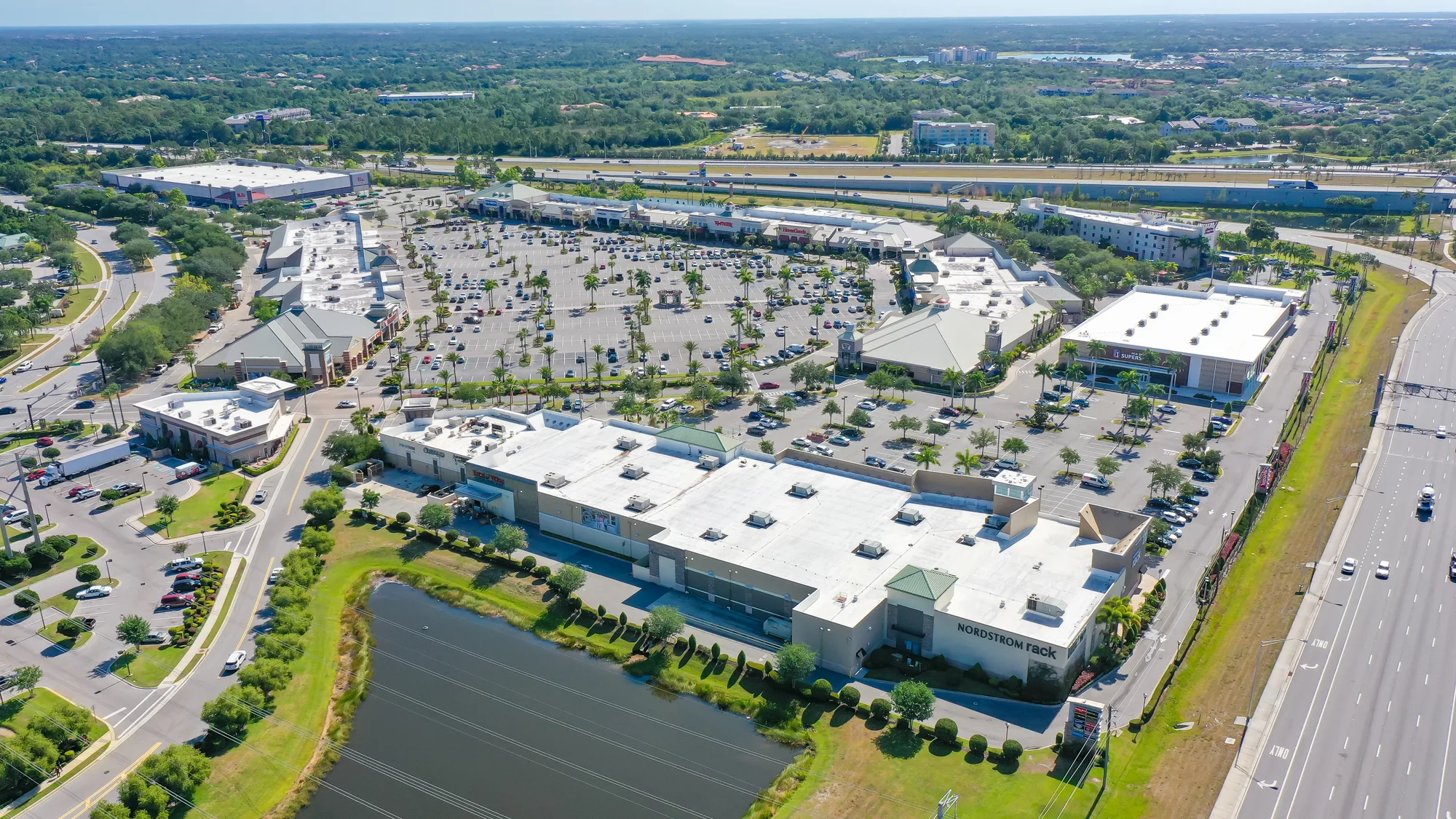 An aerial view of a shopping center featuring a large parking lot filled with vehicles, surrounded by various retail buildings, including a Nordstrom Rack. The image captures the layout of the center with green landscaping and a nearby highway visible in the background.