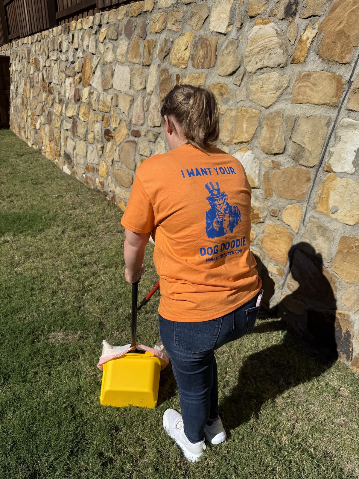 A person wearing an orange t-shirt uses a yellow tool to collect dog waste in a grassy area with a stone wall in the background