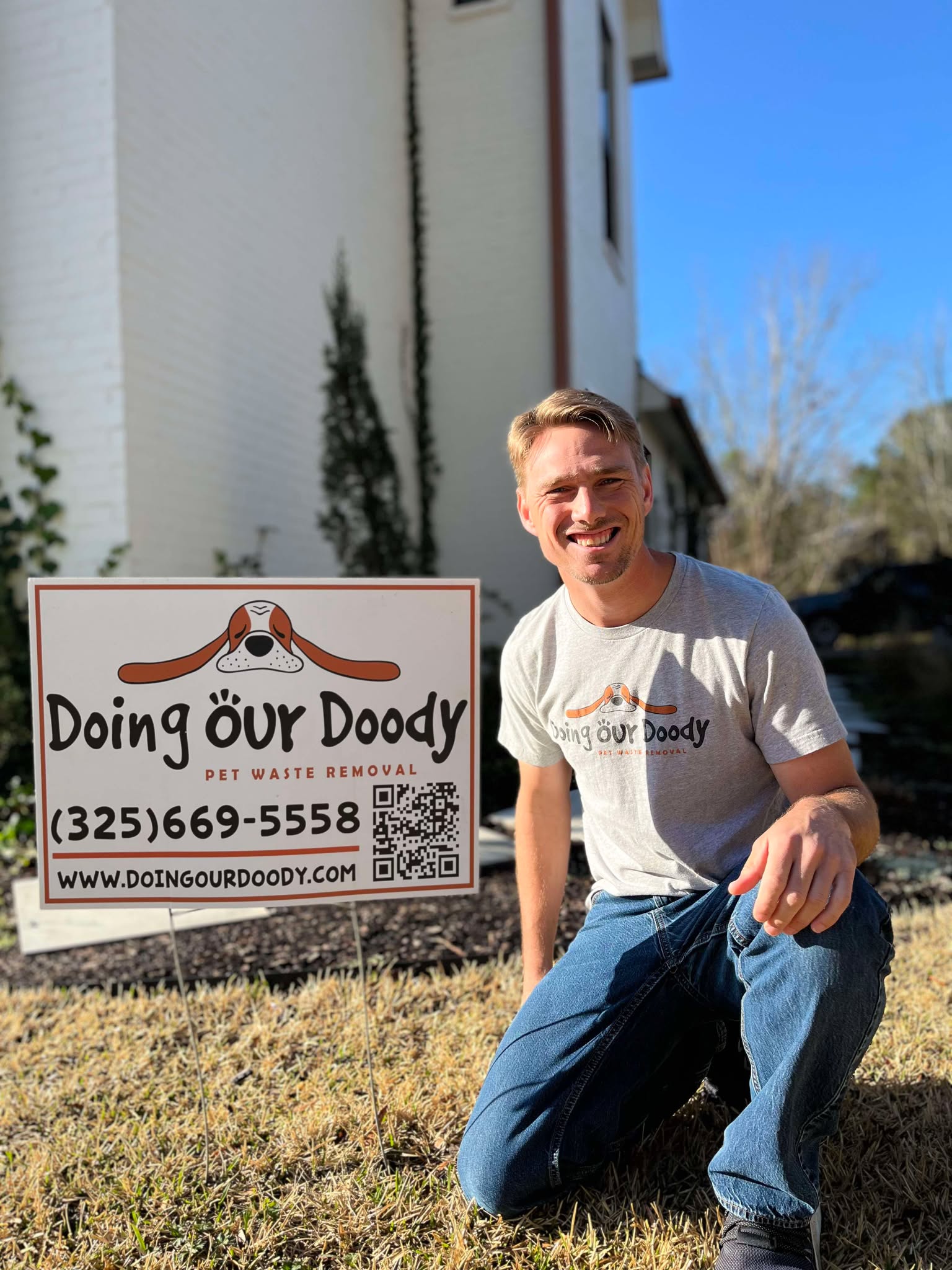 Doing Our Doody owner kneeling beside a yard sign while wearing a branded shirt, with a residential home in the background
