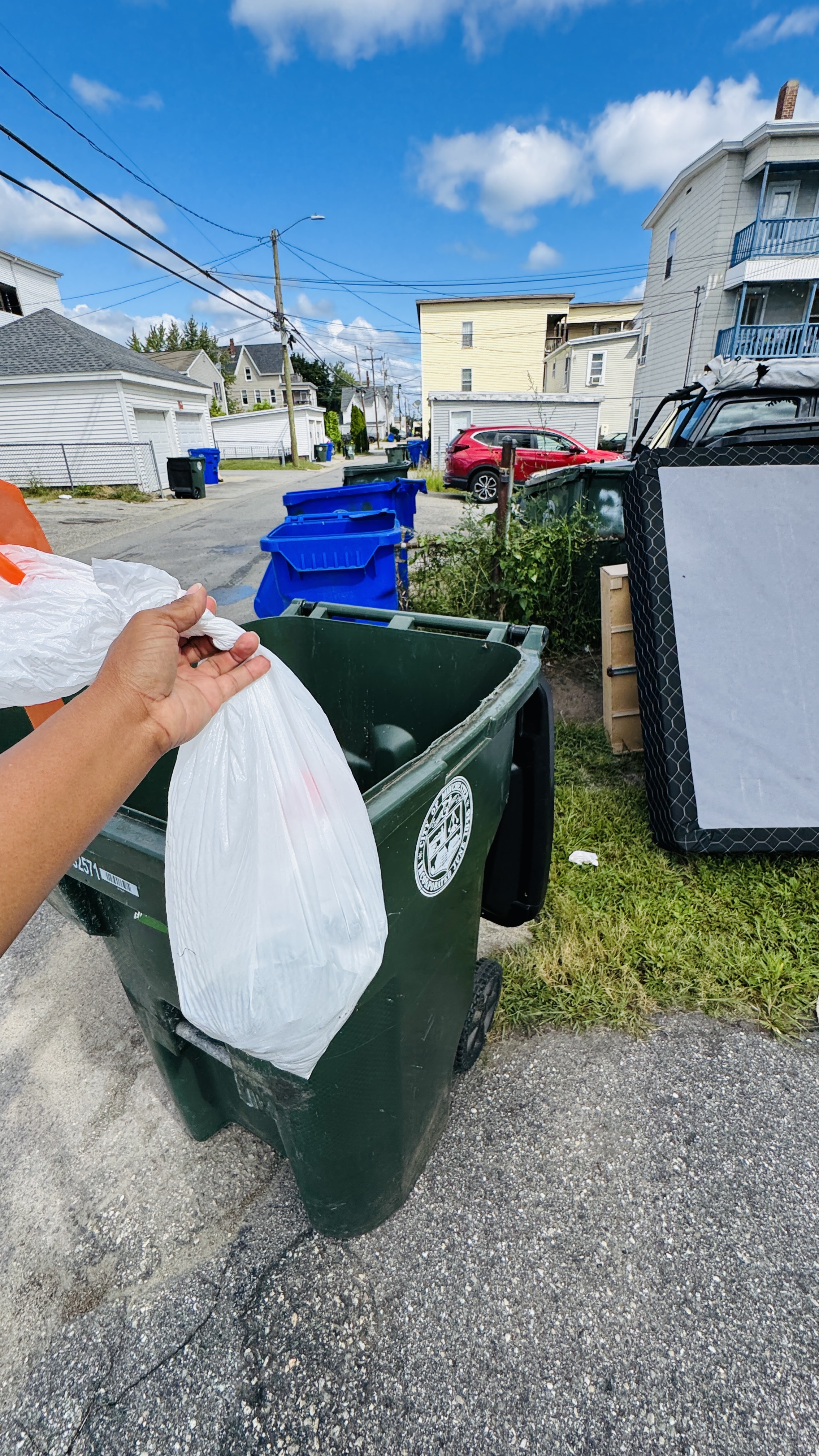 Bagged pet waste being dropped into a green outdoor trash barrel on a residential street