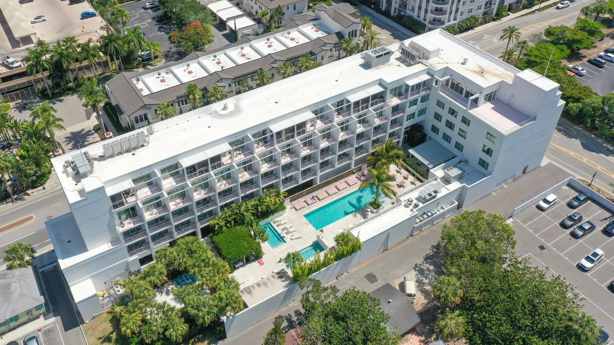 This aerial image shows a modern hotel building with multiple balconies overlooking a swimming pool surrounded by palm trees. The area features landscaped gardens and parking spaces adjacent to the hotel.