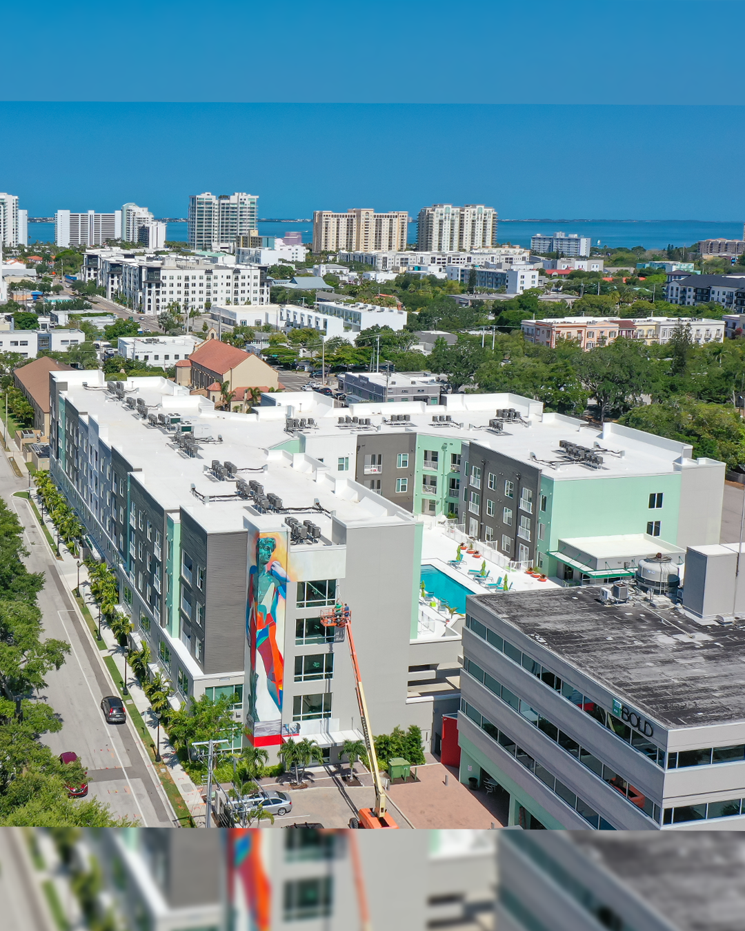 An aerial view of a modern apartment complex featuring a vibrant mural on one side and a swimming pool area. Surrounding the building are other structures and greenery, with a clear view of the city skyline in the distance.