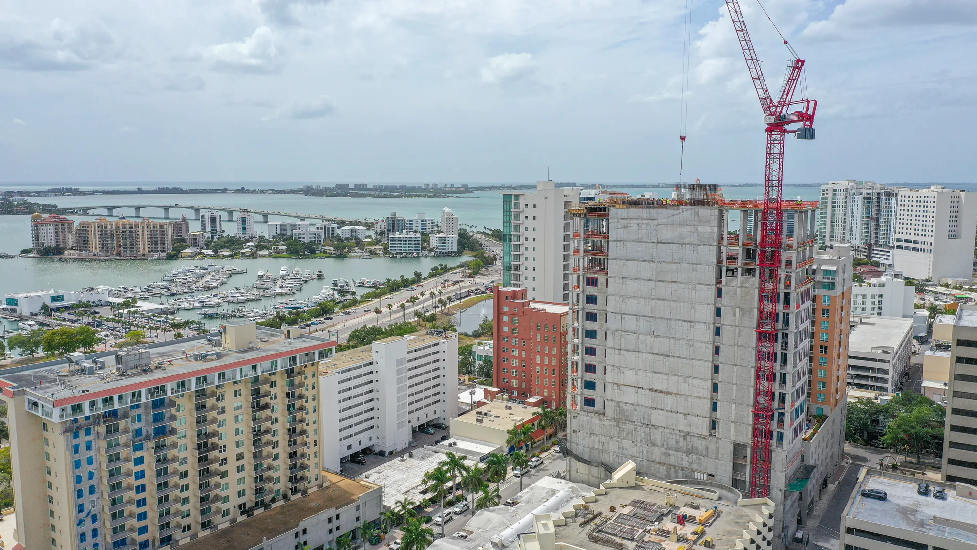 An aerial view of an ongoing construction site in a city, featuring a tall building with a red crane. The background showcases a marina with boats, various residential buildings, and water, emphasizing the urban landscape.