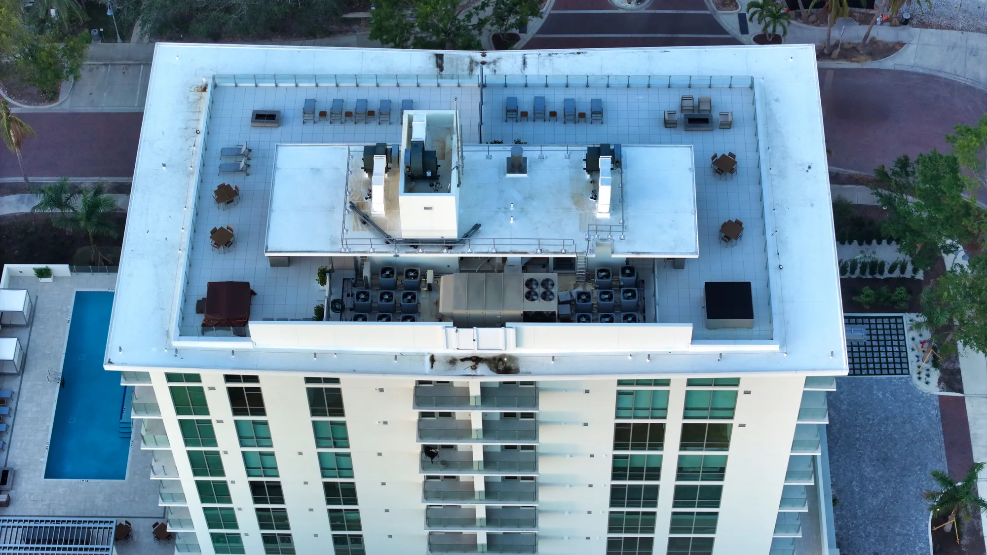 The image shows the rooftop of a modern residential building with a flat, white surface. Featuring several outdoor seating areas, air conditioning units, and a clear view of the surrounding area, this rooftop space offers amenities for residents.