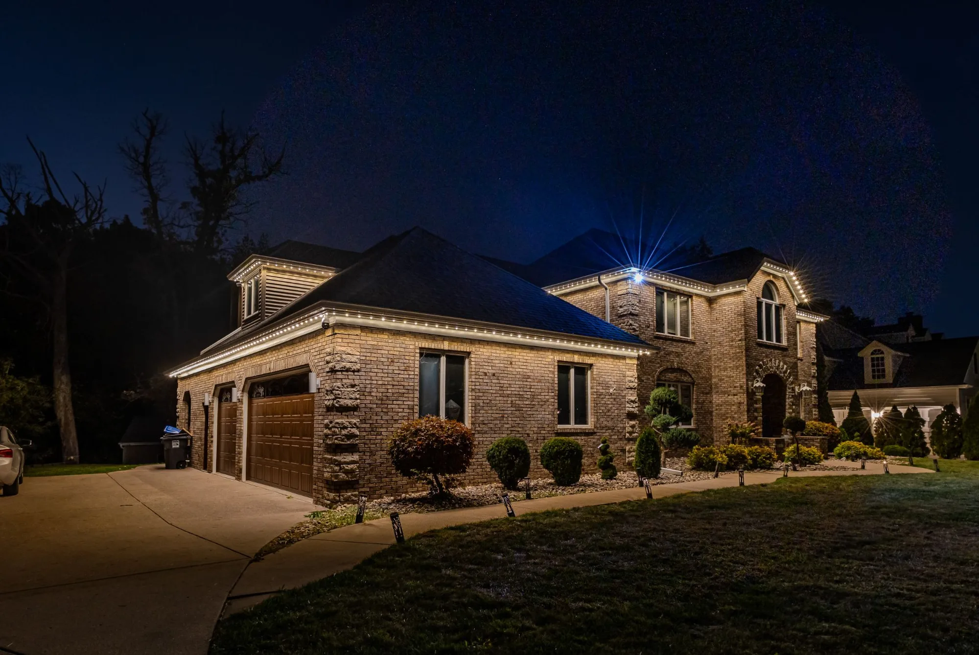 Large brick home at night with permanent roofline lighting set to warm white, tracing the eaves and gables, with pathway lights along the front walk.