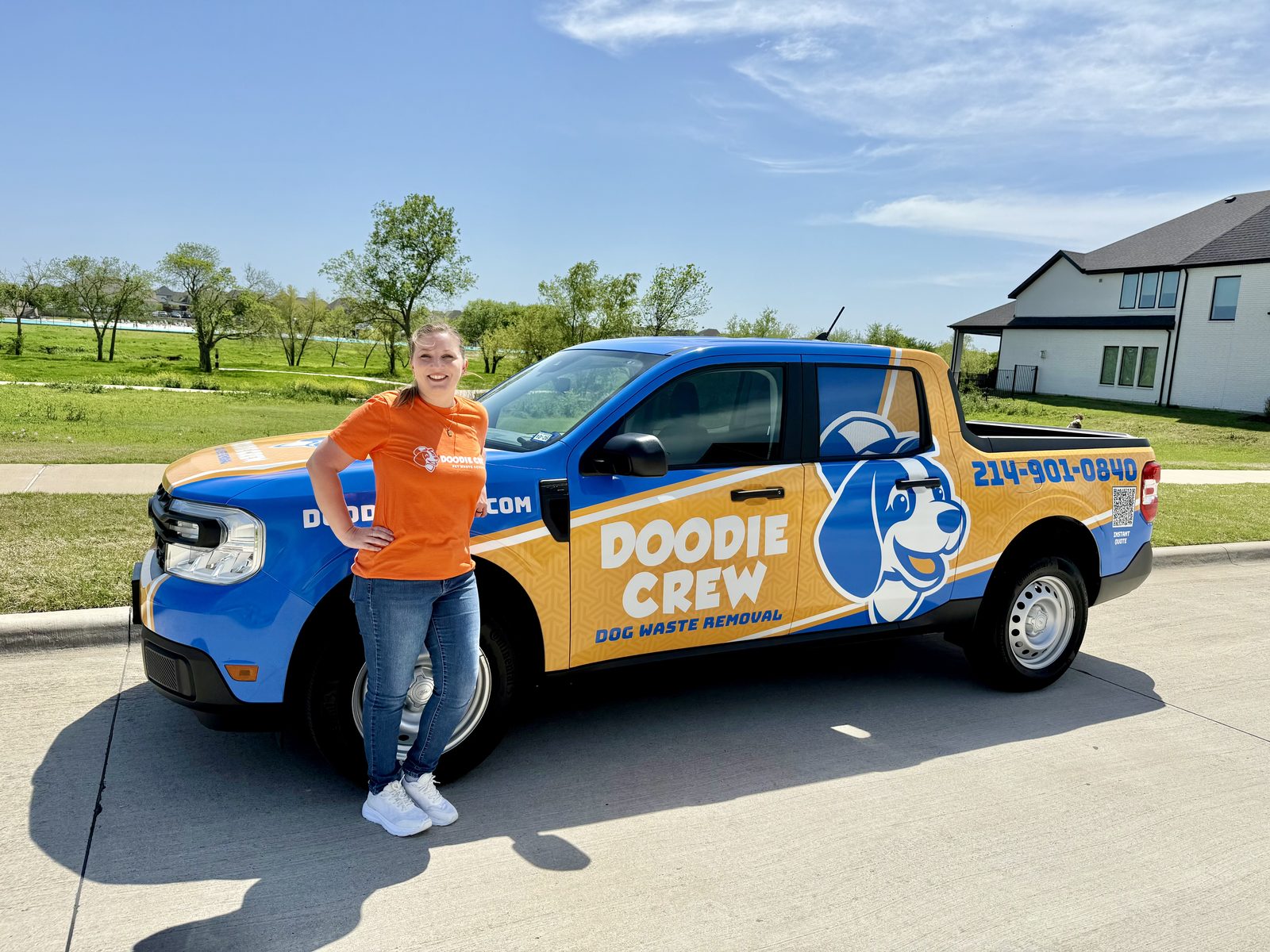 A woman in an orange t-shirt stands beside the Doodie Crew Dog Waste Removal truck in front of a modern house with a green landscape