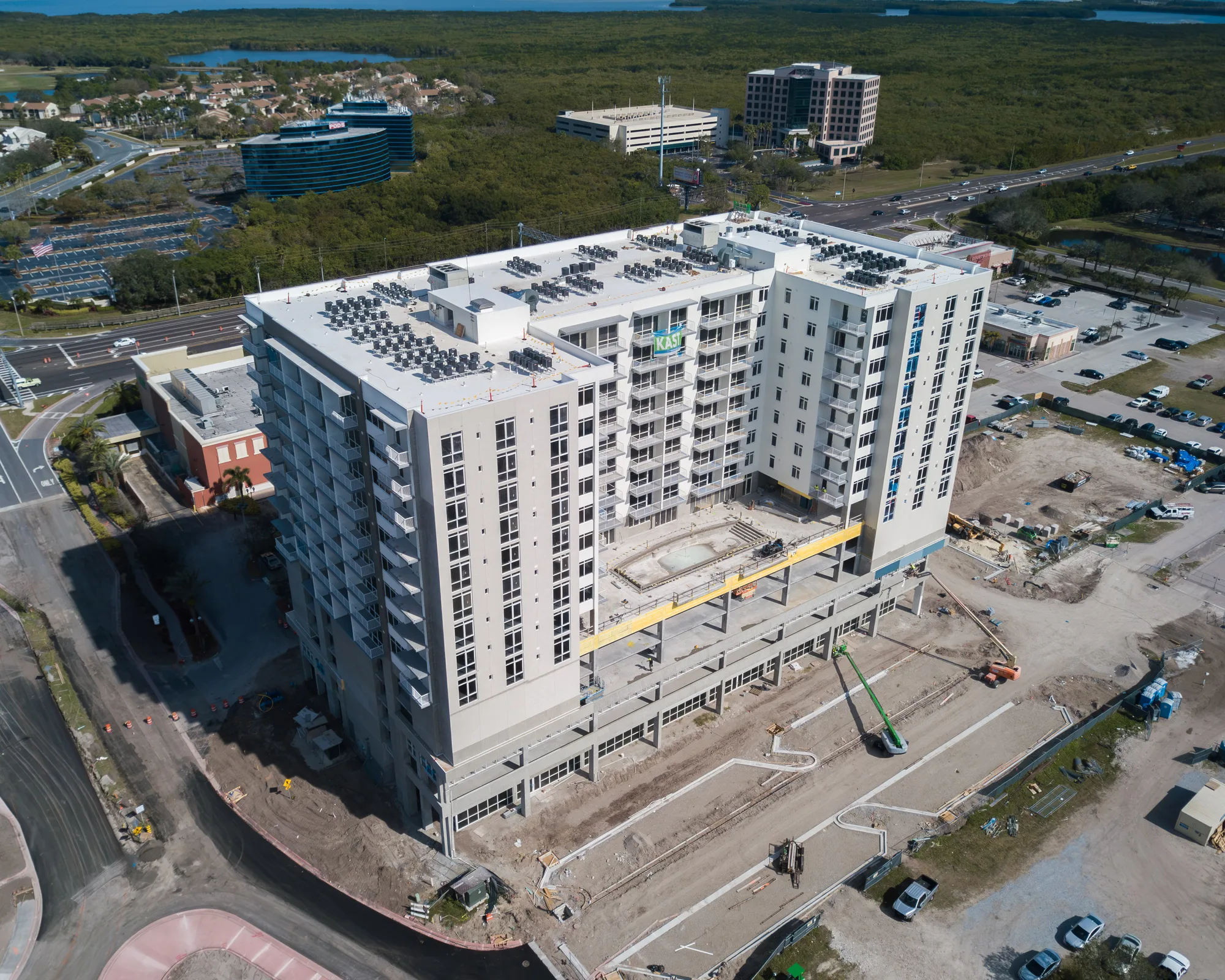 Aerial of The Collection Residences in Sarasota, a white multi-storey residential building among the downtown low-rise blocks, with the Gulf beyond.